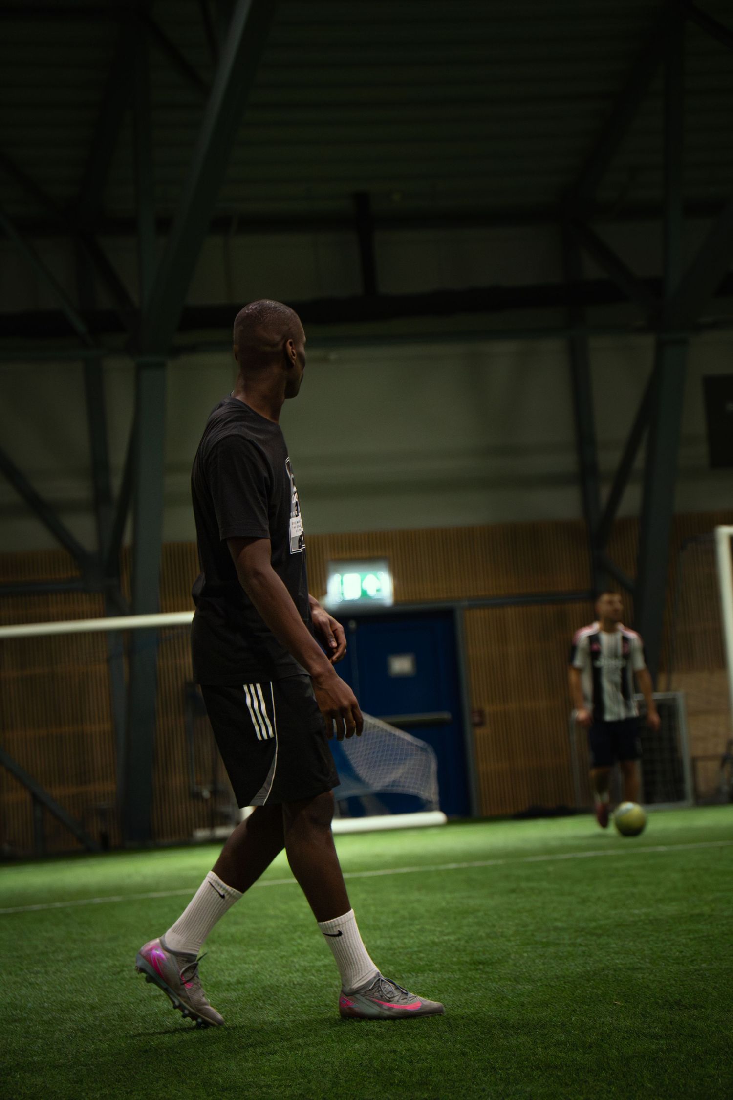 Two men playing indoor soccer on artificial turf in a sports hall, one in black Adidas kit, one in striped jersey.
