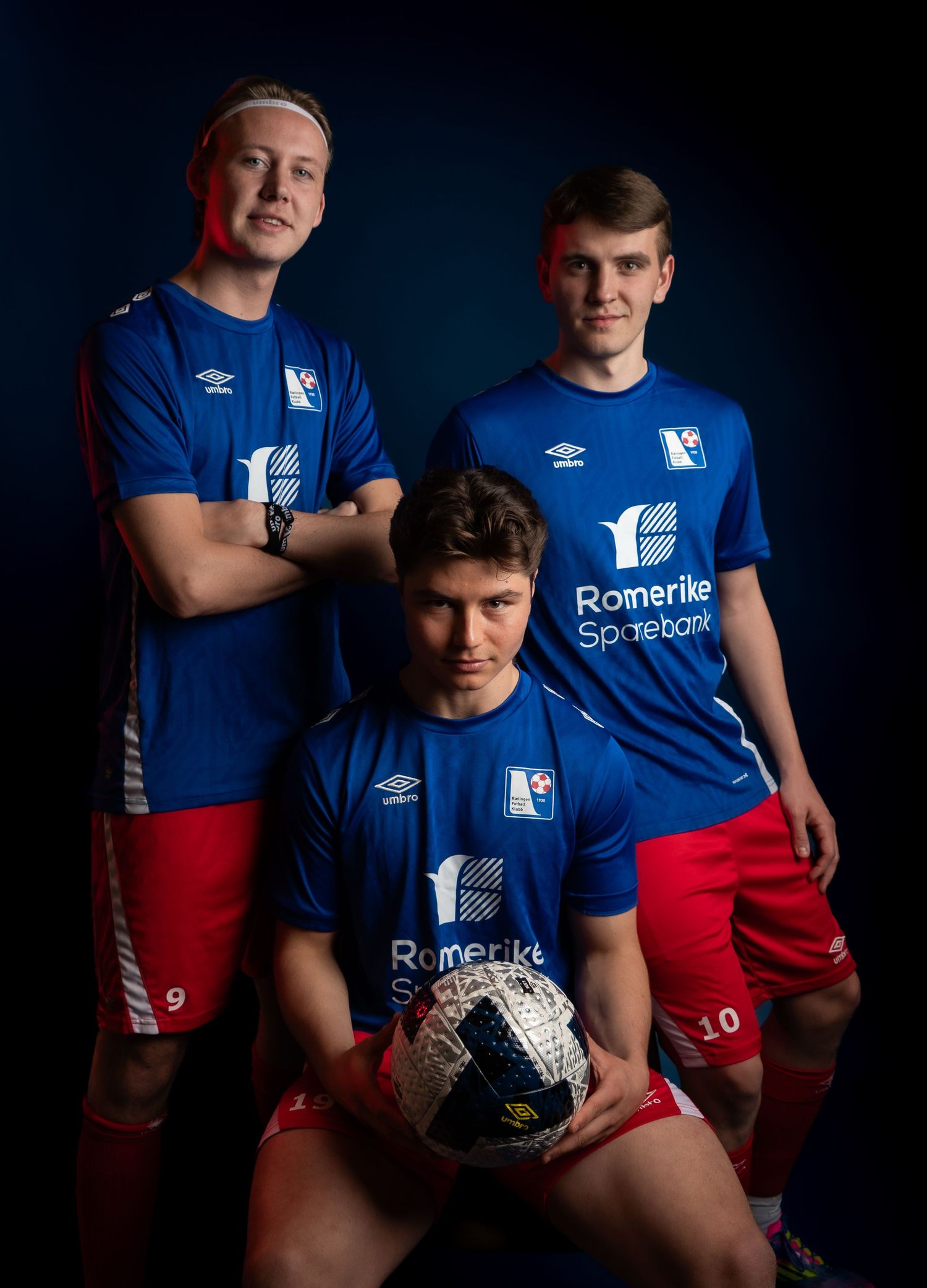 Team players in blue and red uniforms pose with handball in dramatic studio lighting.