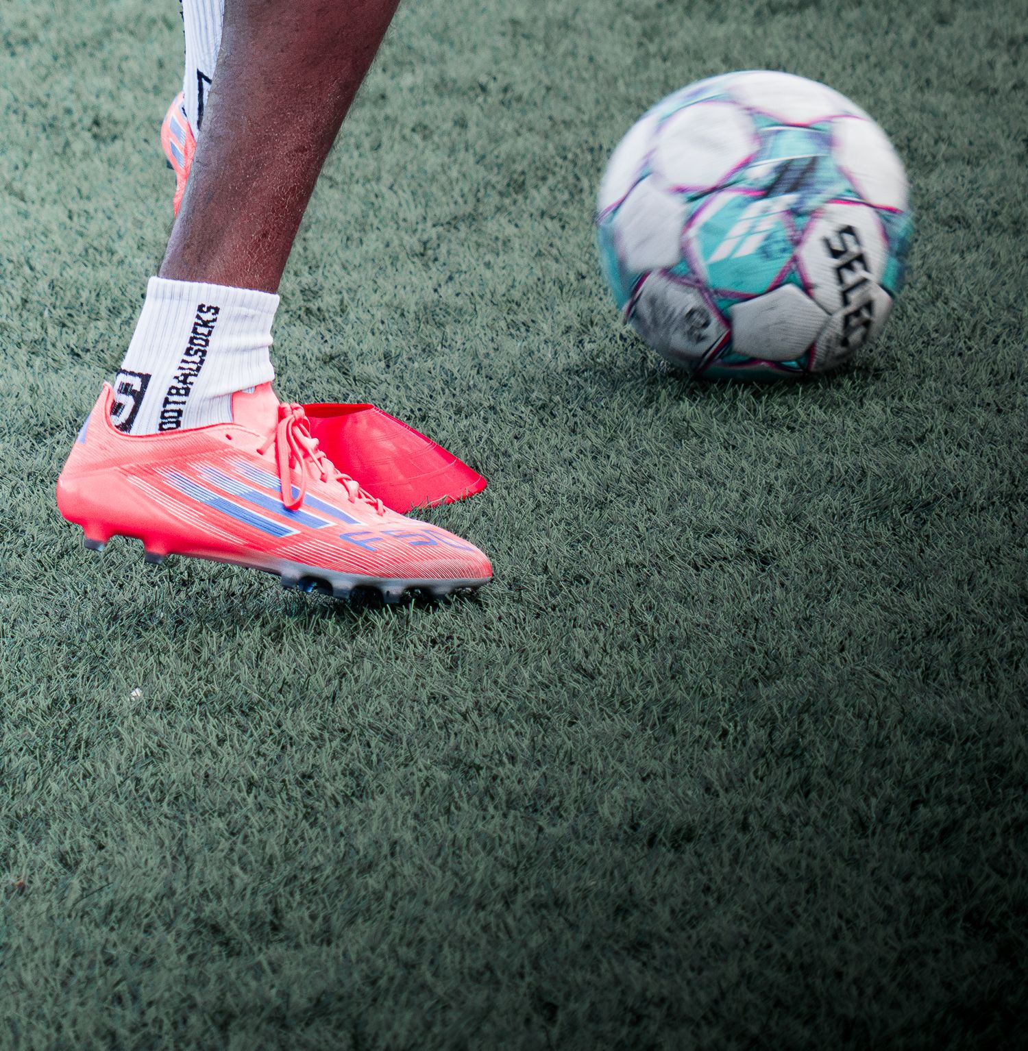 Close-up of a soccer player wearing bright coral cleats on artificial turf beside a Select soccer ball.