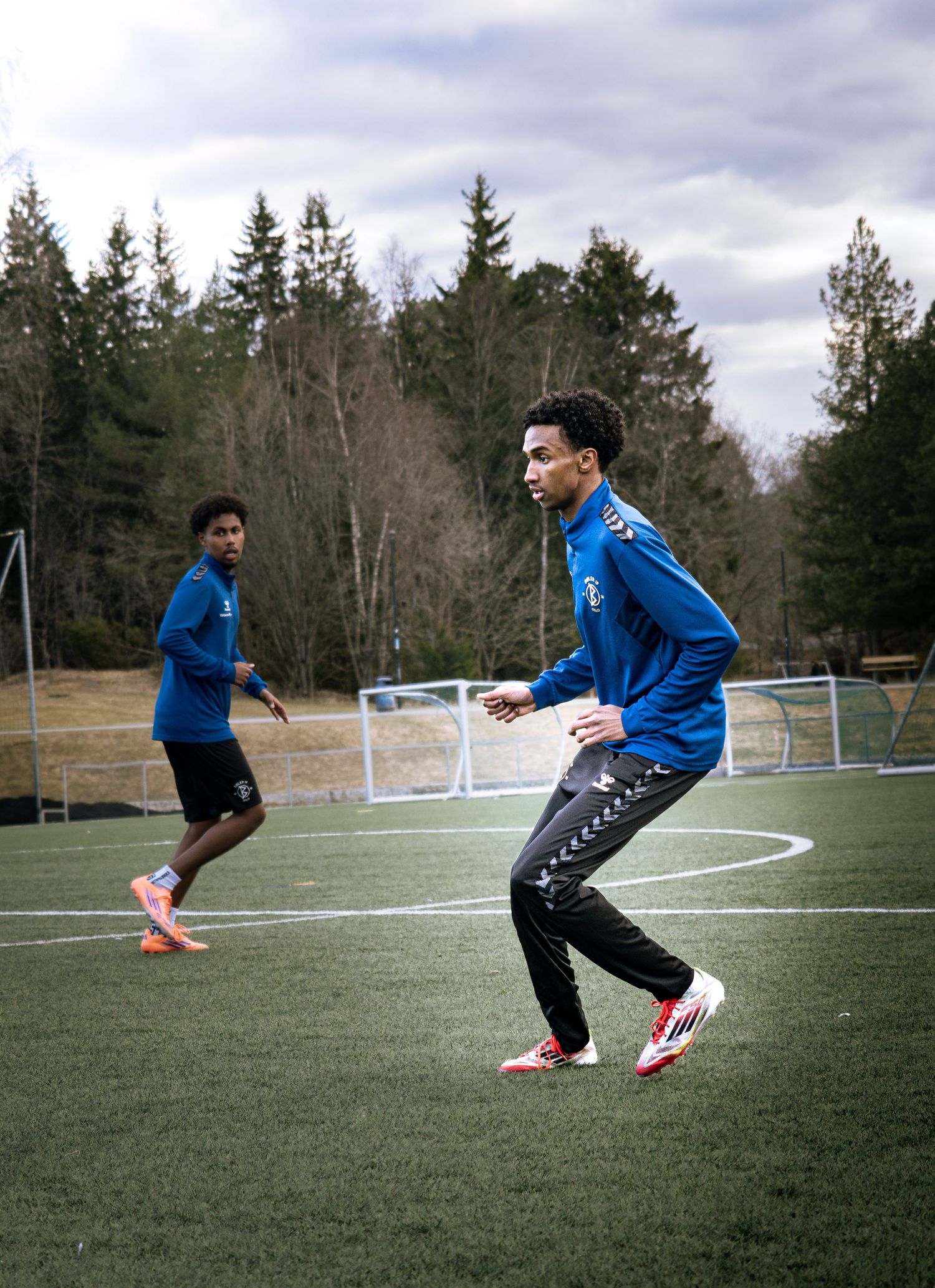 Two soccer players in blue Hummel uniforms training on an outdoor artificial turf field surrounded by trees.