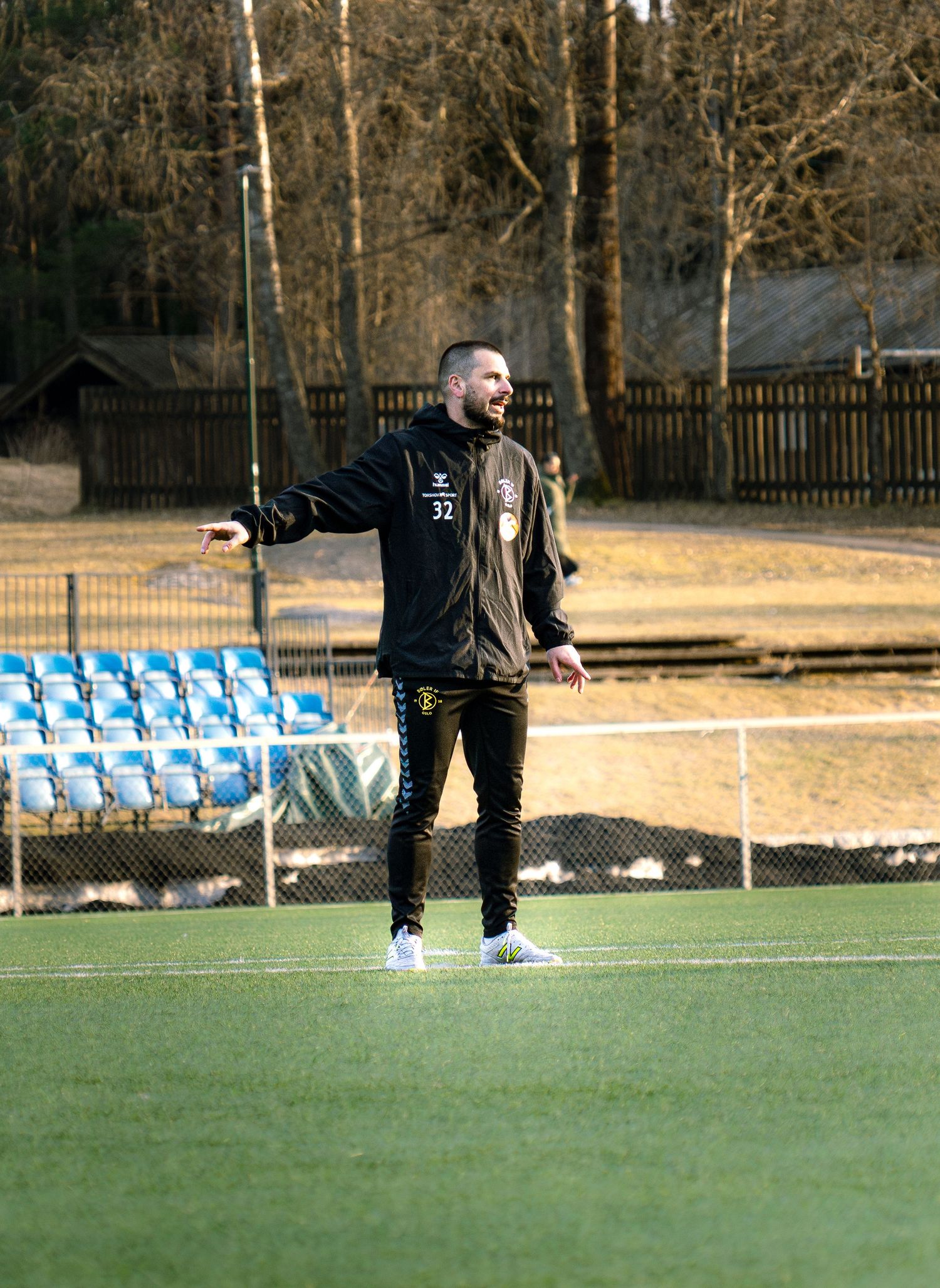 A football coach in black tracksuit number 32 gestures on a green pitch beside empty blue stadium seats.