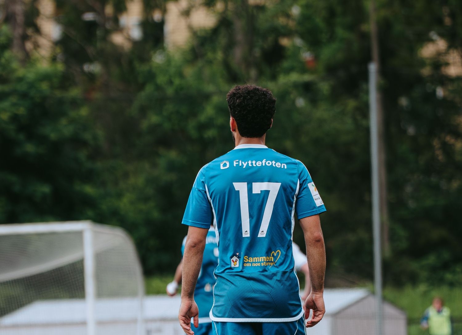 Soccer player wearing blue number 17 jersey with Flyttefotan sponsor stands on field with green trees background.