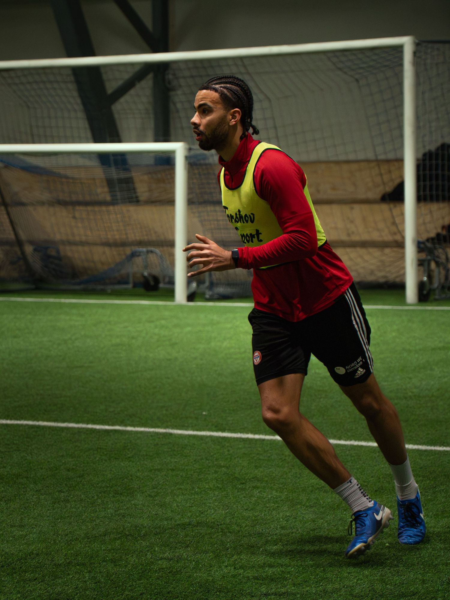 Male soccer player in red jersey and yellow vest running on indoor turf field during training session.