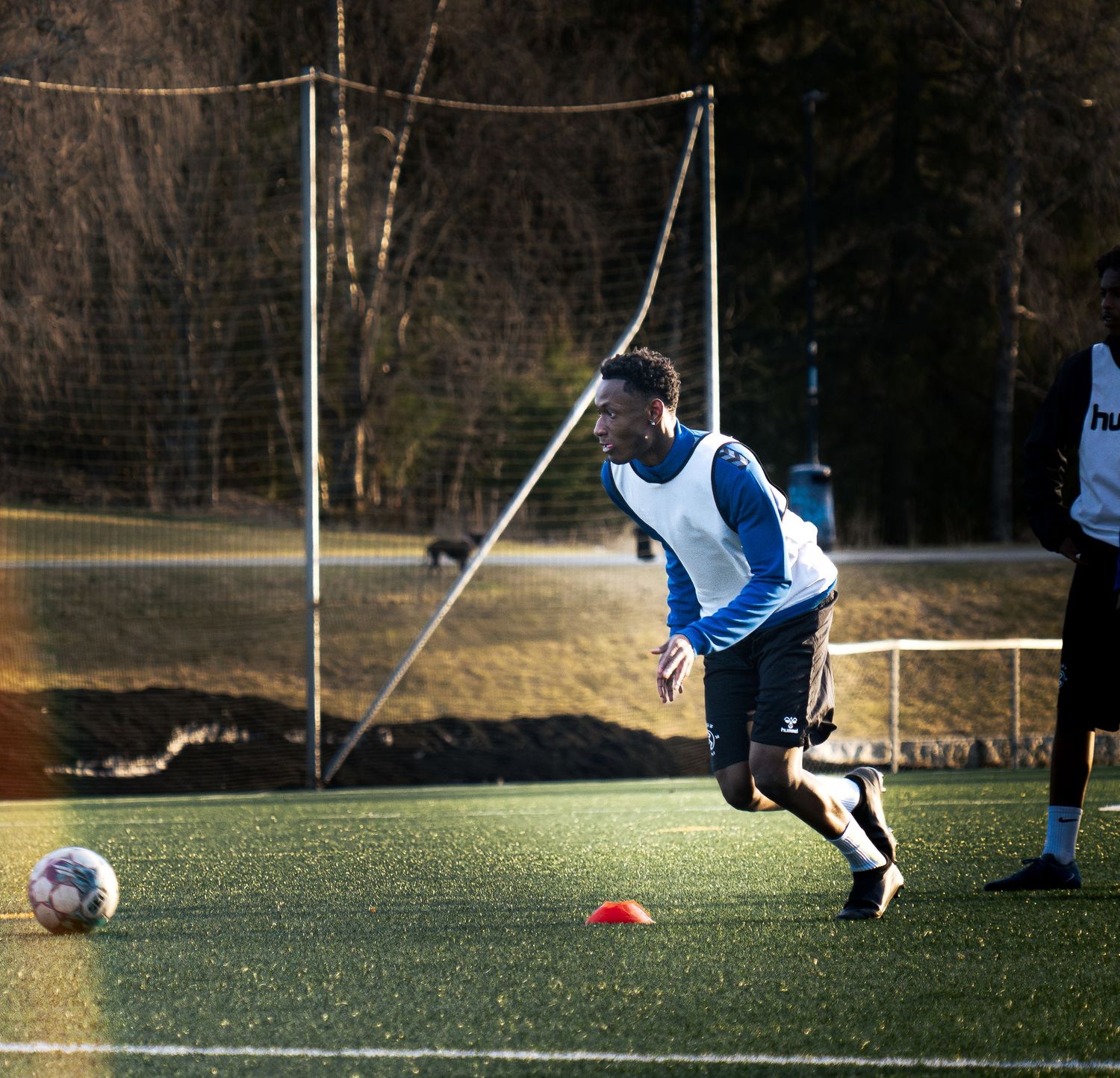 Soccer player in blue vest dribbles ball on green turf field during outdoor training session on a sunny day.