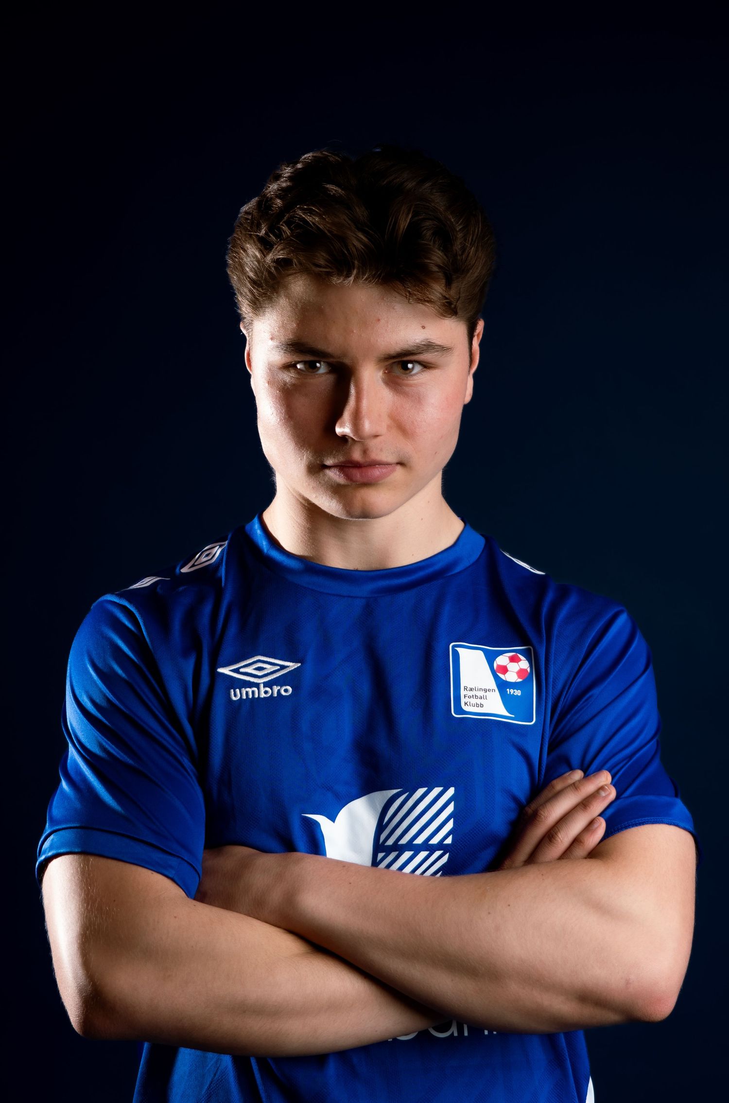 Sports player in blue uniform poses confidently against dark background with crossed arms.