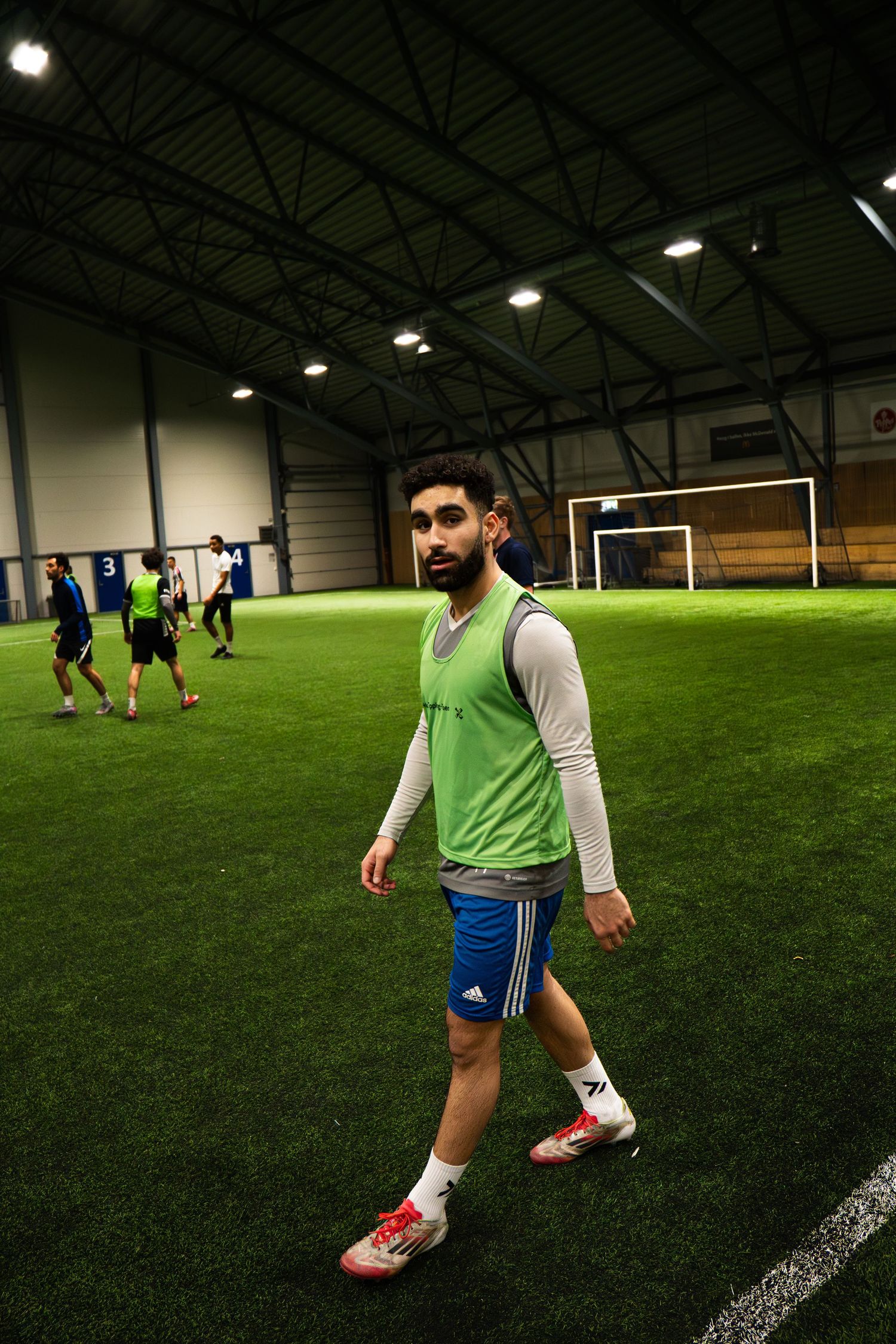 Young male soccer player in green vest and blue Adidas shorts walking on indoor turf field during practice.