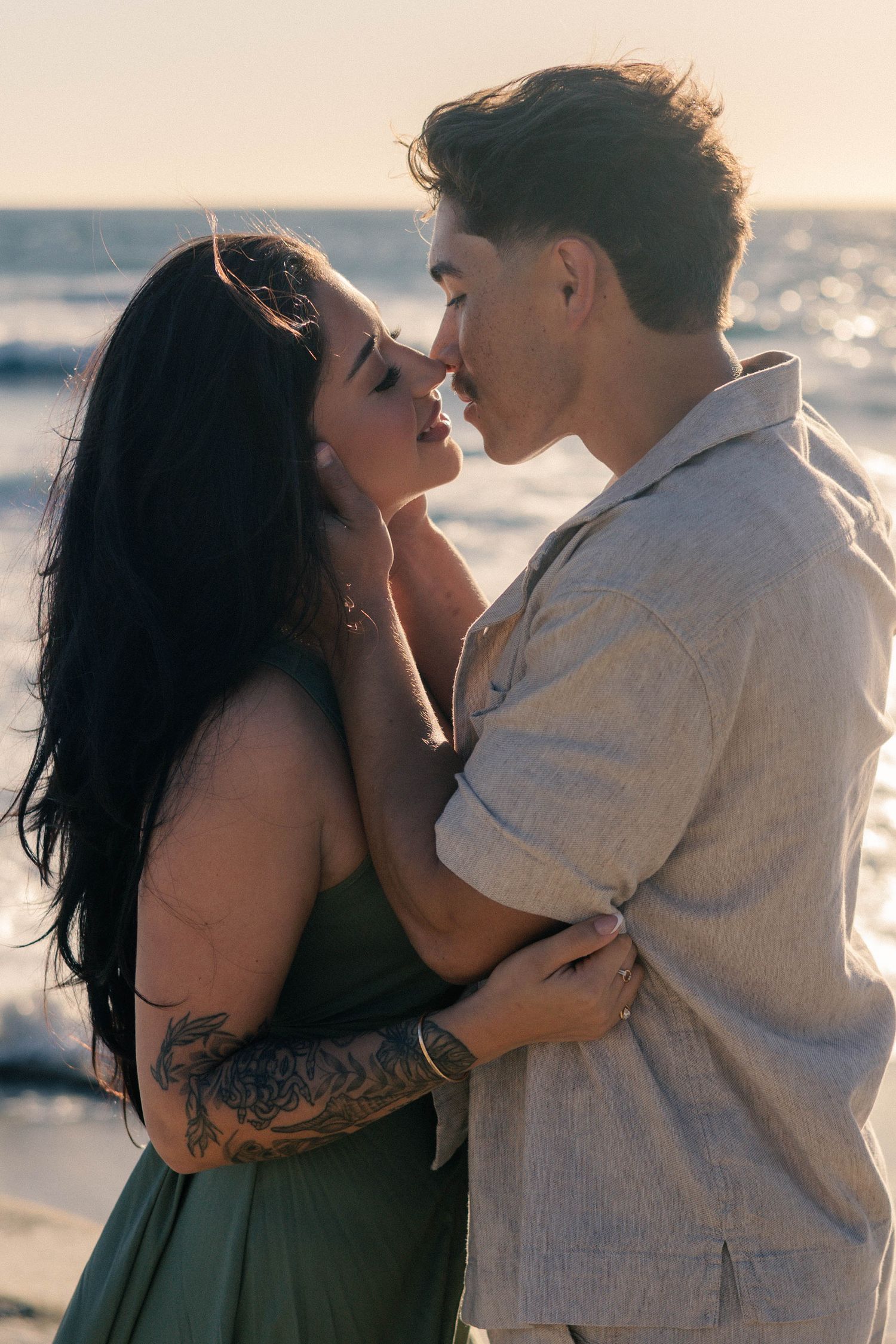 Couple embracing and nearly kissing on a beach at sunset, woman wearing green dress with tattoos.