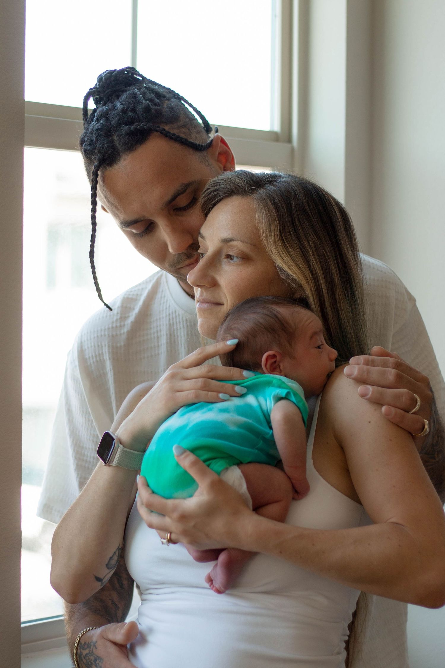 New parents share a tender moment, father kissing mothers head as she cradles their newborn baby in a green onesie.