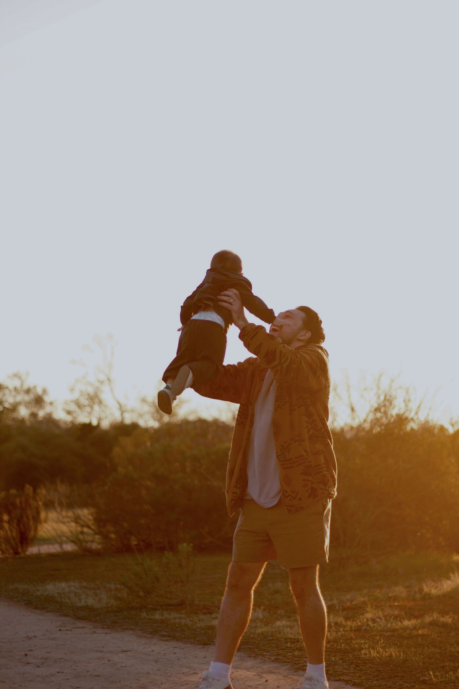 Father joyfully lifting baby into the air at golden hour in a park setting.