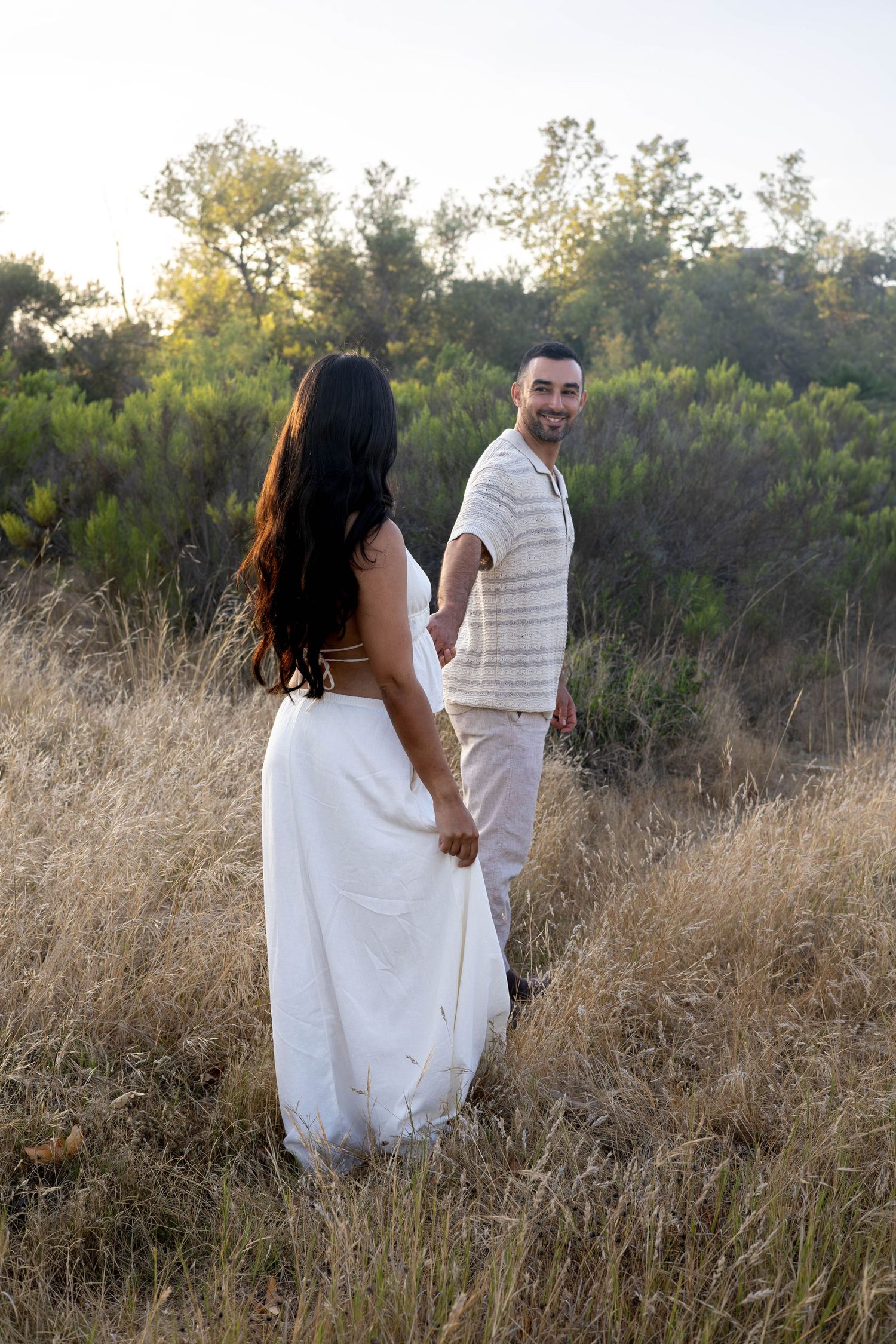 Couple holding hands in golden field, woman in white dress, man in cream outfit, lush green trees backdrop.
