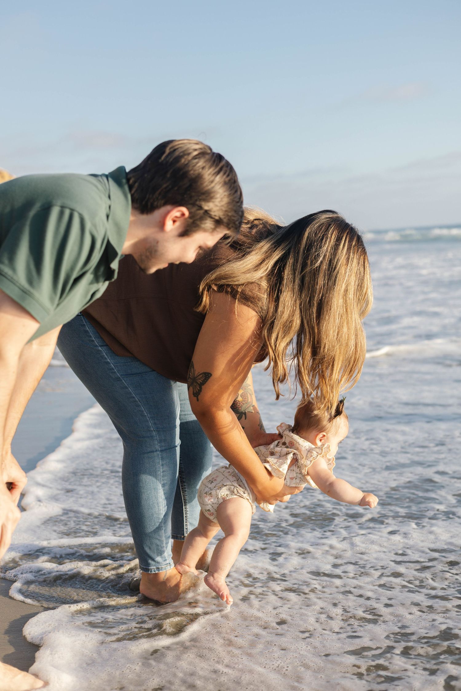 Young couple helping their baby touch ocean waves at the beach on a sunny day.