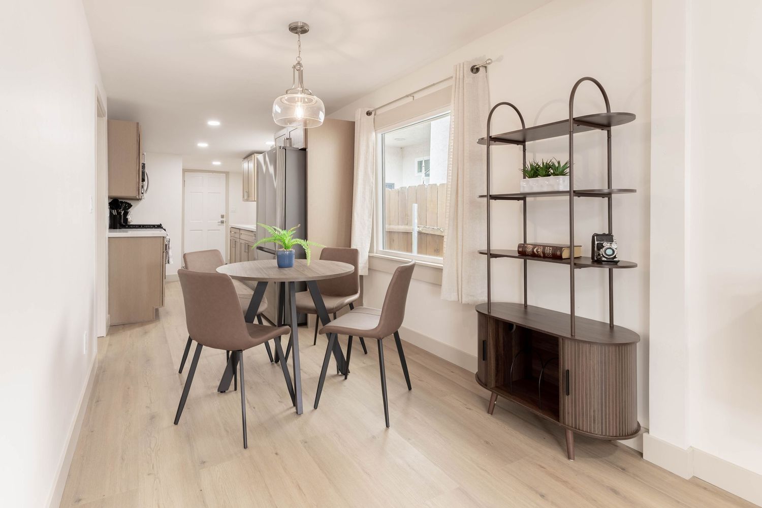 Modern dining area with round table, taupe chairs, chandelier, light hardwood floors, and metal shelving unit.