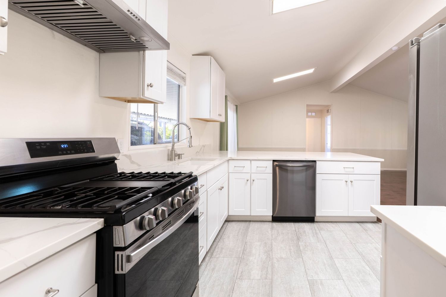 Bright kitchen featuring gas range, black dishwasher, white shaker cabinets, vaulted ceiling, and hardwood floors.