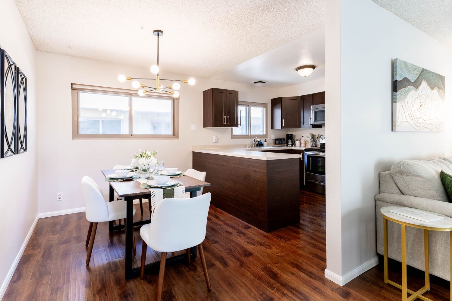 Modern dining area with white chairs and dark wood table, open to kitchen with dark cabinets and hardwood floors.