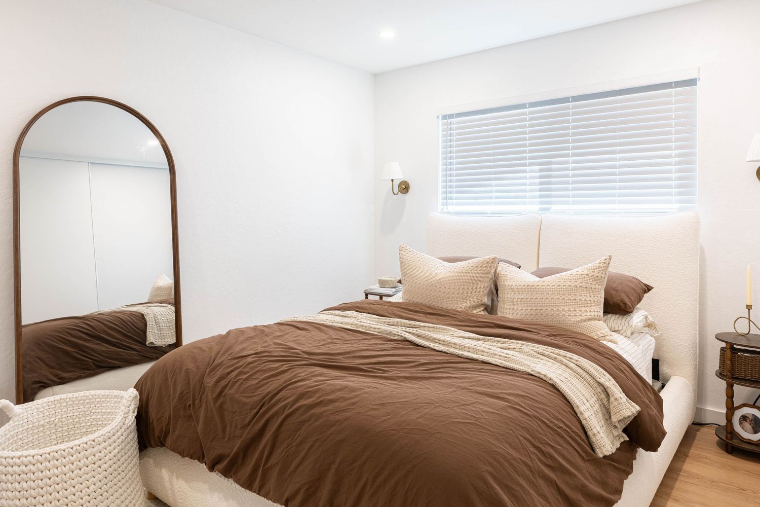 Modern bedroom with brown duvet, beige pillows, arched floor mirror, and white walls with natural light.
