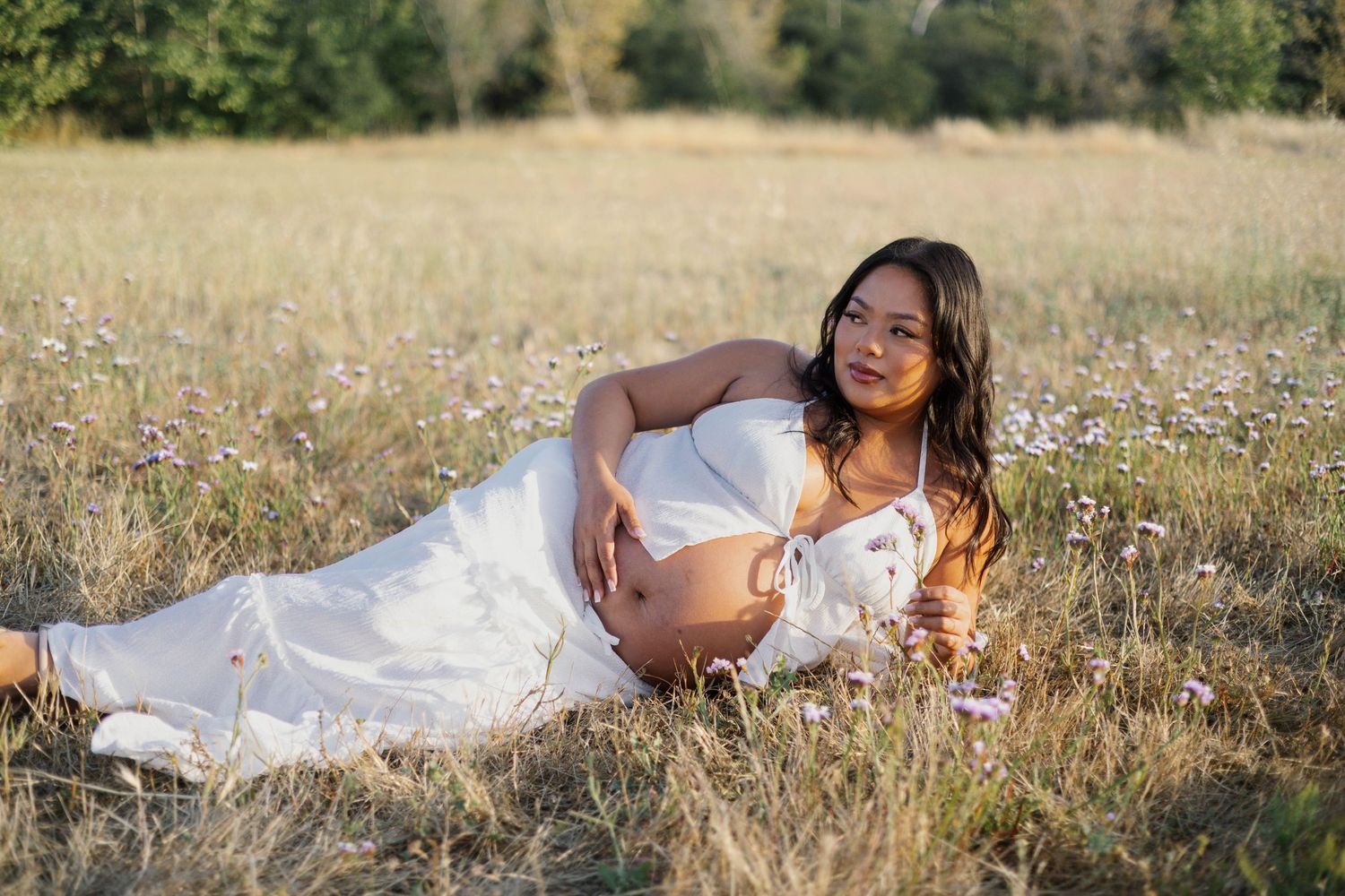 Pregnant woman in white dress lying in a wildflower field in golden sunlight.