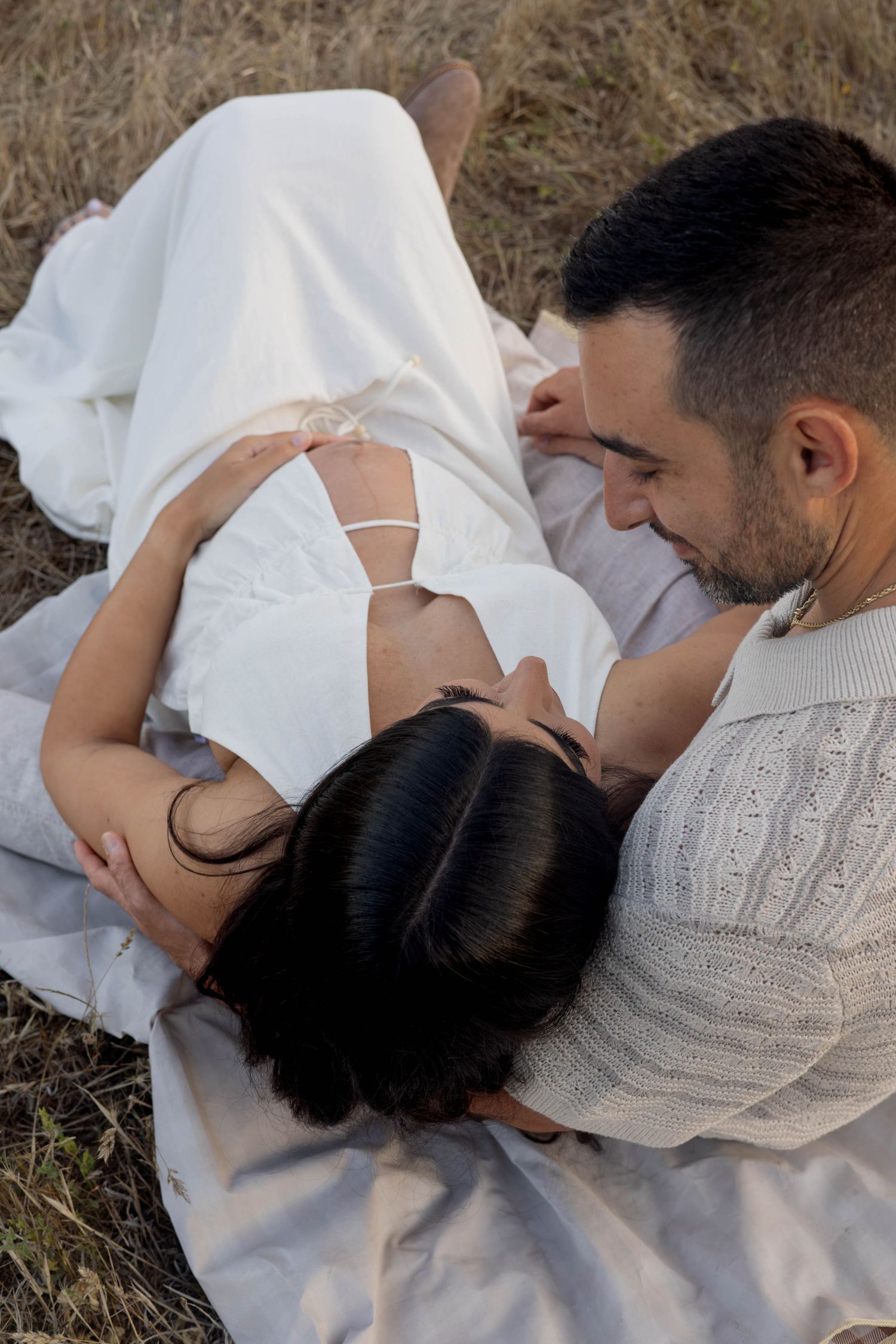 Couple lying on blanket in field, pregnant woman in white dress with partner smiling at her baby bump.