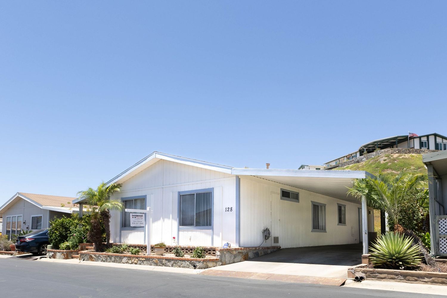White single-story mobile home with palm trees and landscaping on a sunny residential street in a mobile home park.
