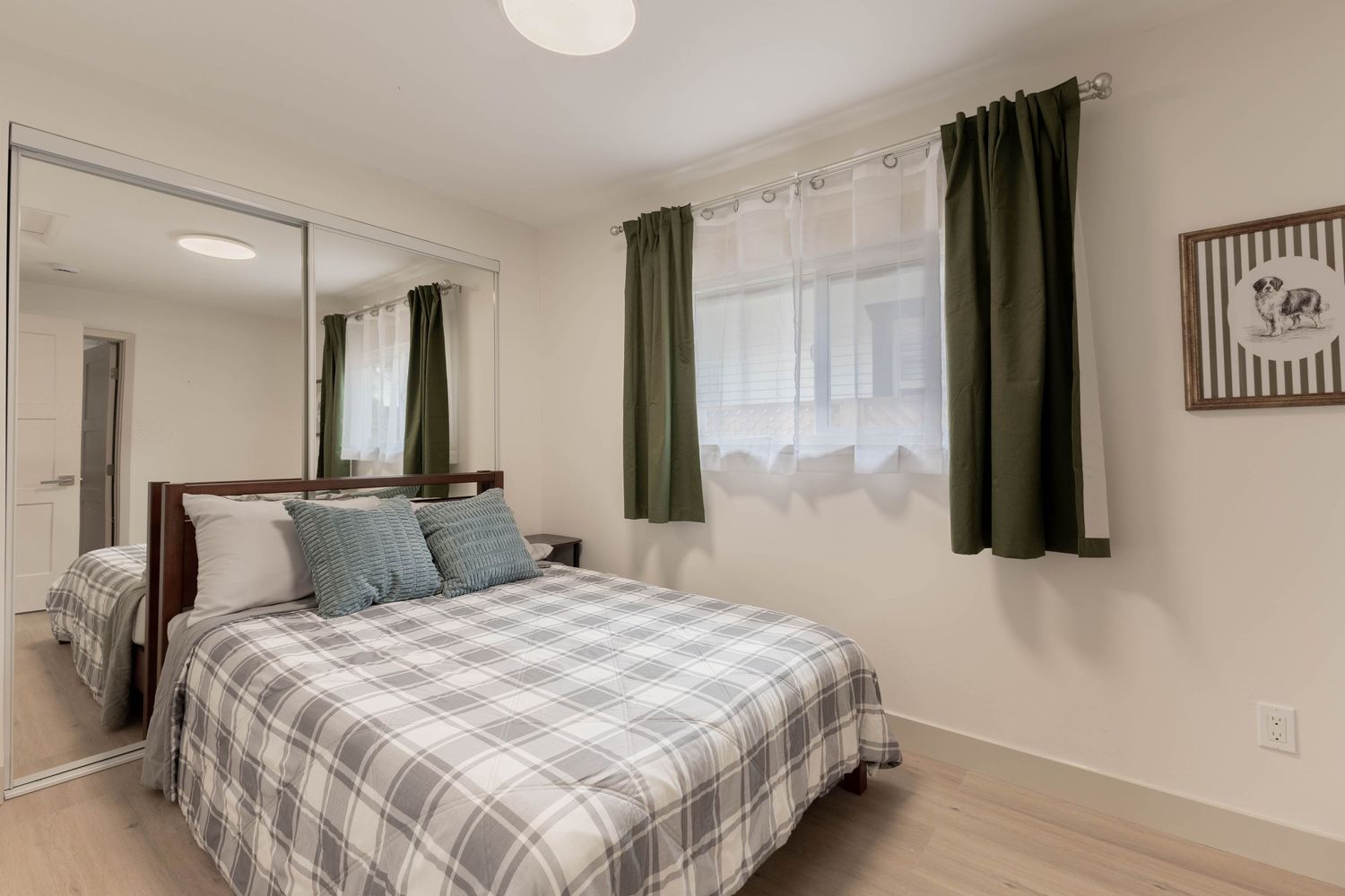 Cozy bedroom with a plaid bedspread, mirrored closet, green curtains, and light hardwood floors.