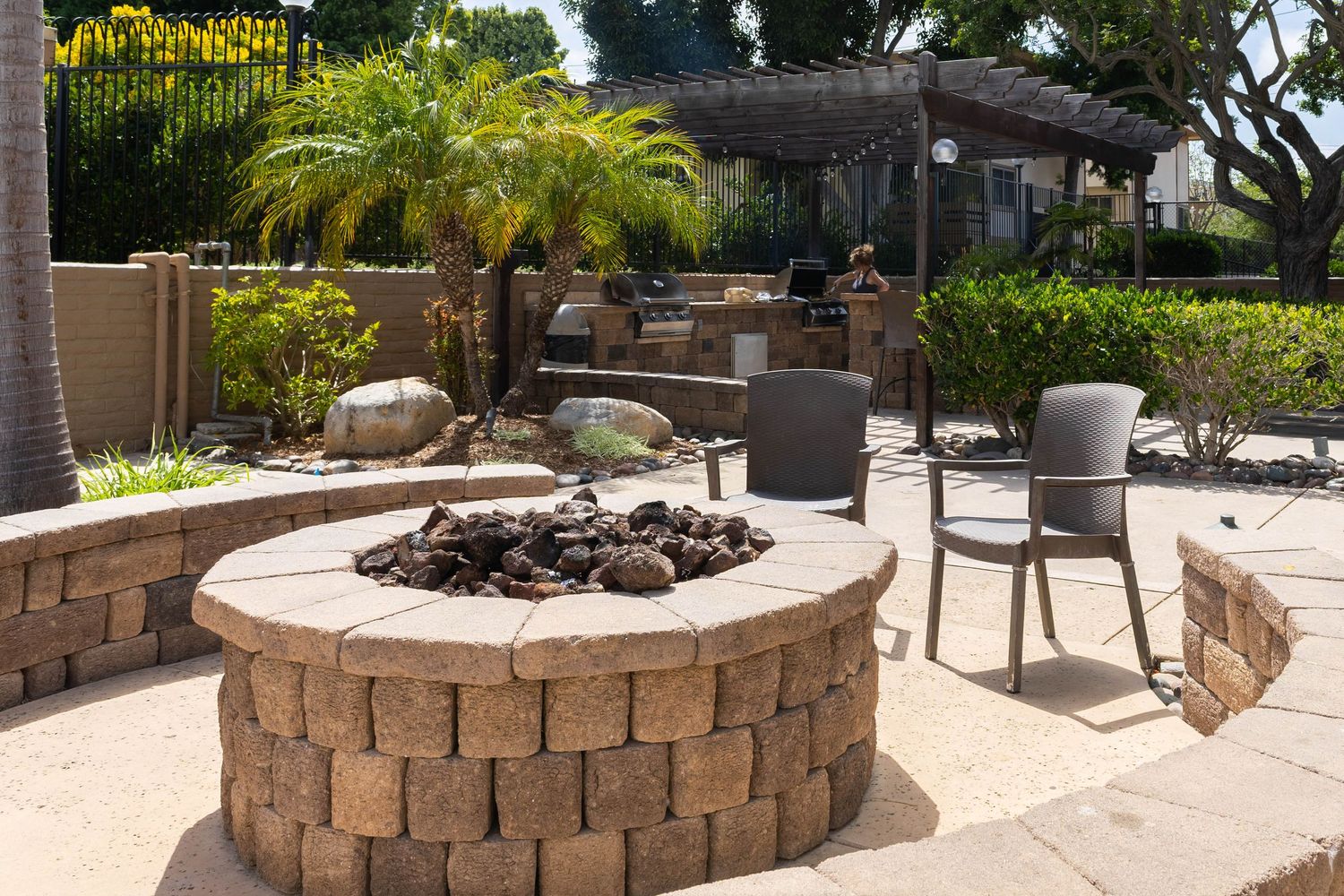 Outdoor patio with stone fire pit, gray chairs, pergola, and lush tropical landscaping on a sunny day.