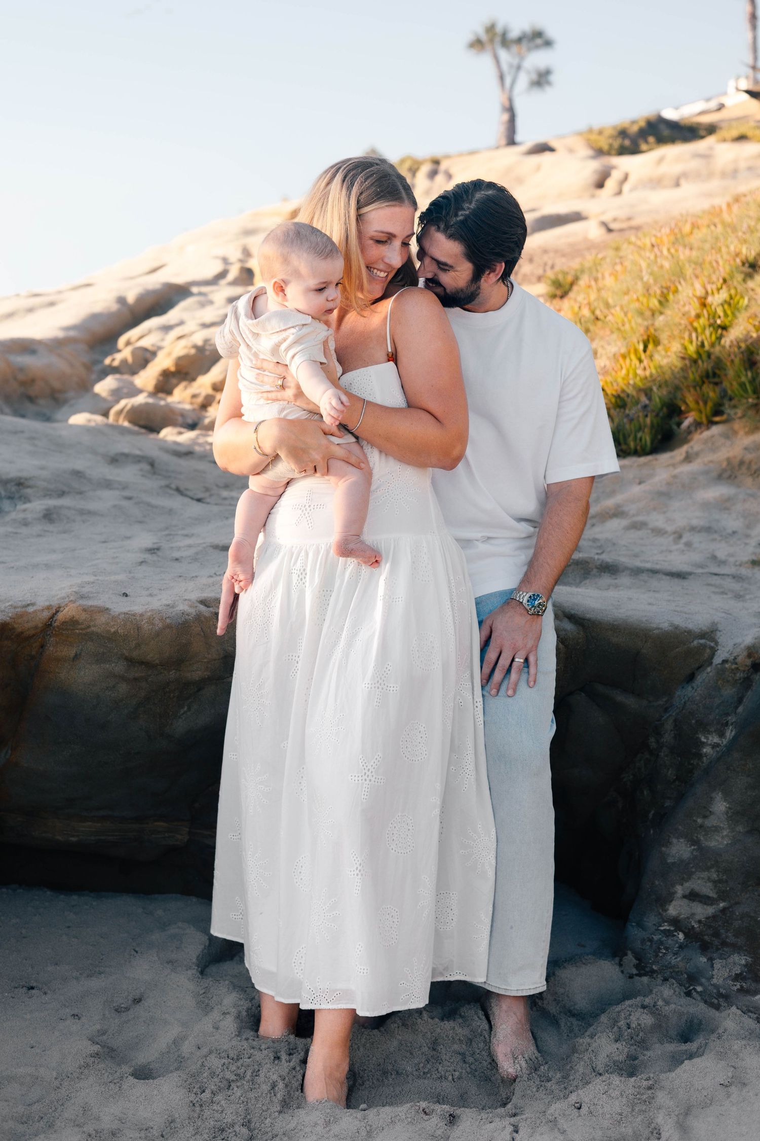 A young family with a baby poses on a sandy beach near rocks, wearing white and light-colored outfits.