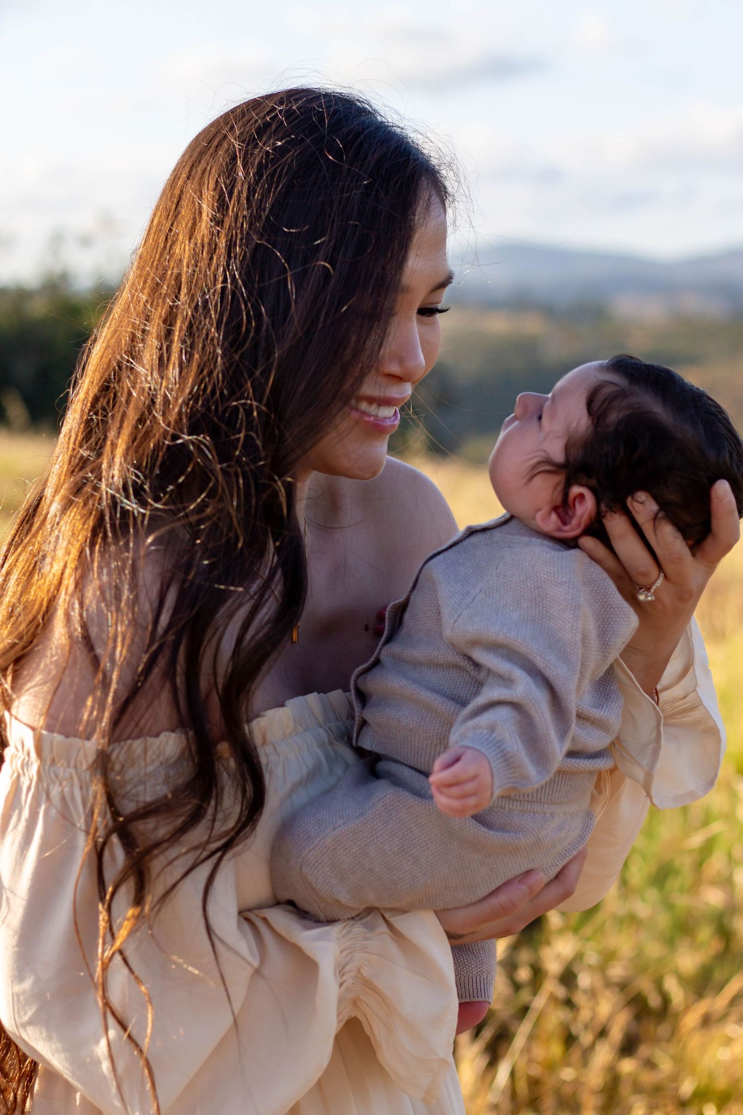 Smiling dark-haired mother in cream dress tenderly holds newborn baby in sunlit field.