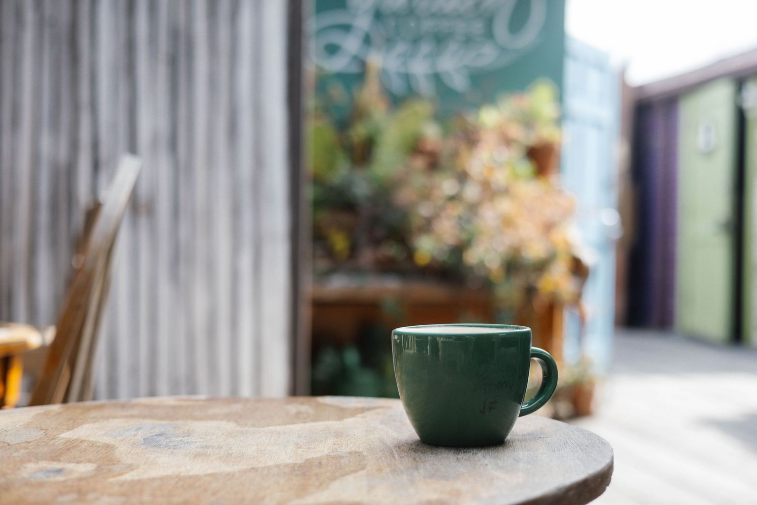 Green ceramic coffee mug on a rustic wooden table outside a cozy cafe with lush greenery in the background.