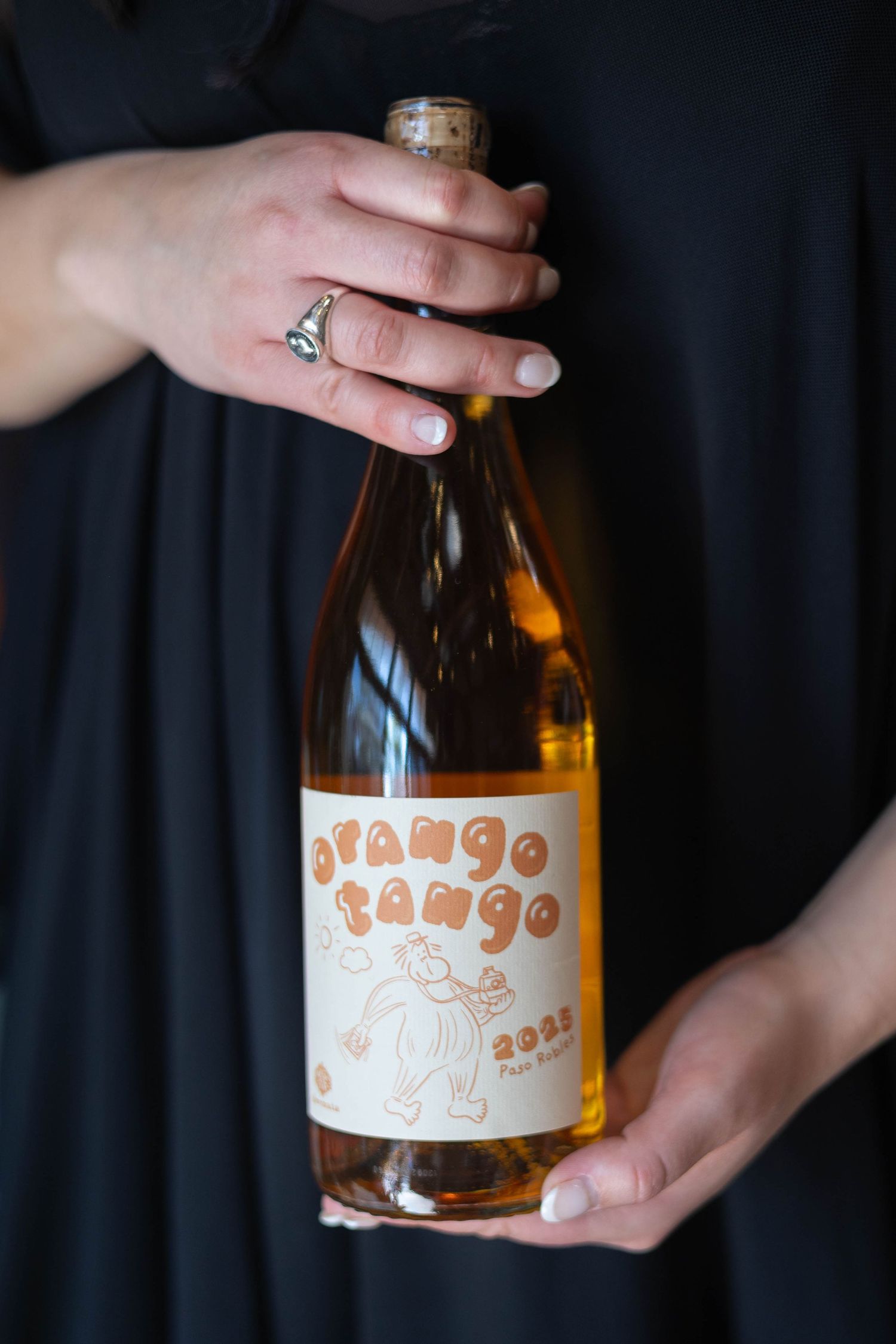 Hands holding a bottle of Orange Tango 2022 Petite Sirah wine with a playful orange label.