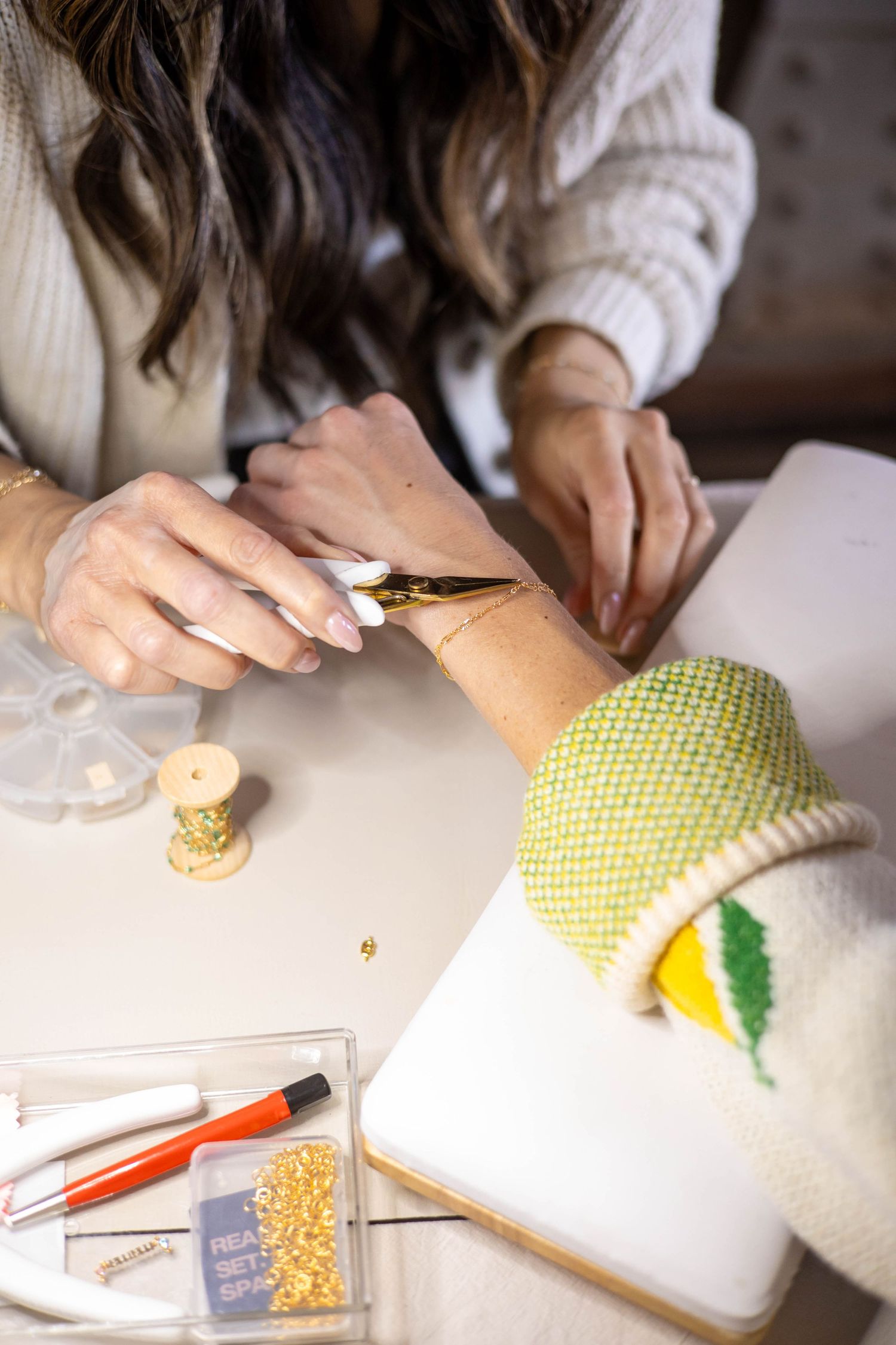 A woman decorates a white cake with gold paint using a fine brush, with beads and tools on the table.