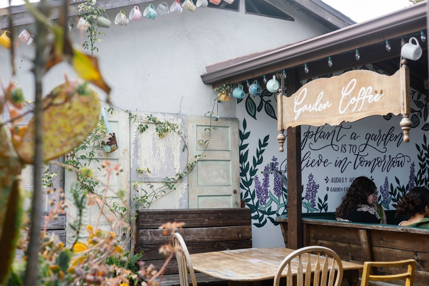 Cozy garden coffee shop patio with floral mural and string lights overhead.