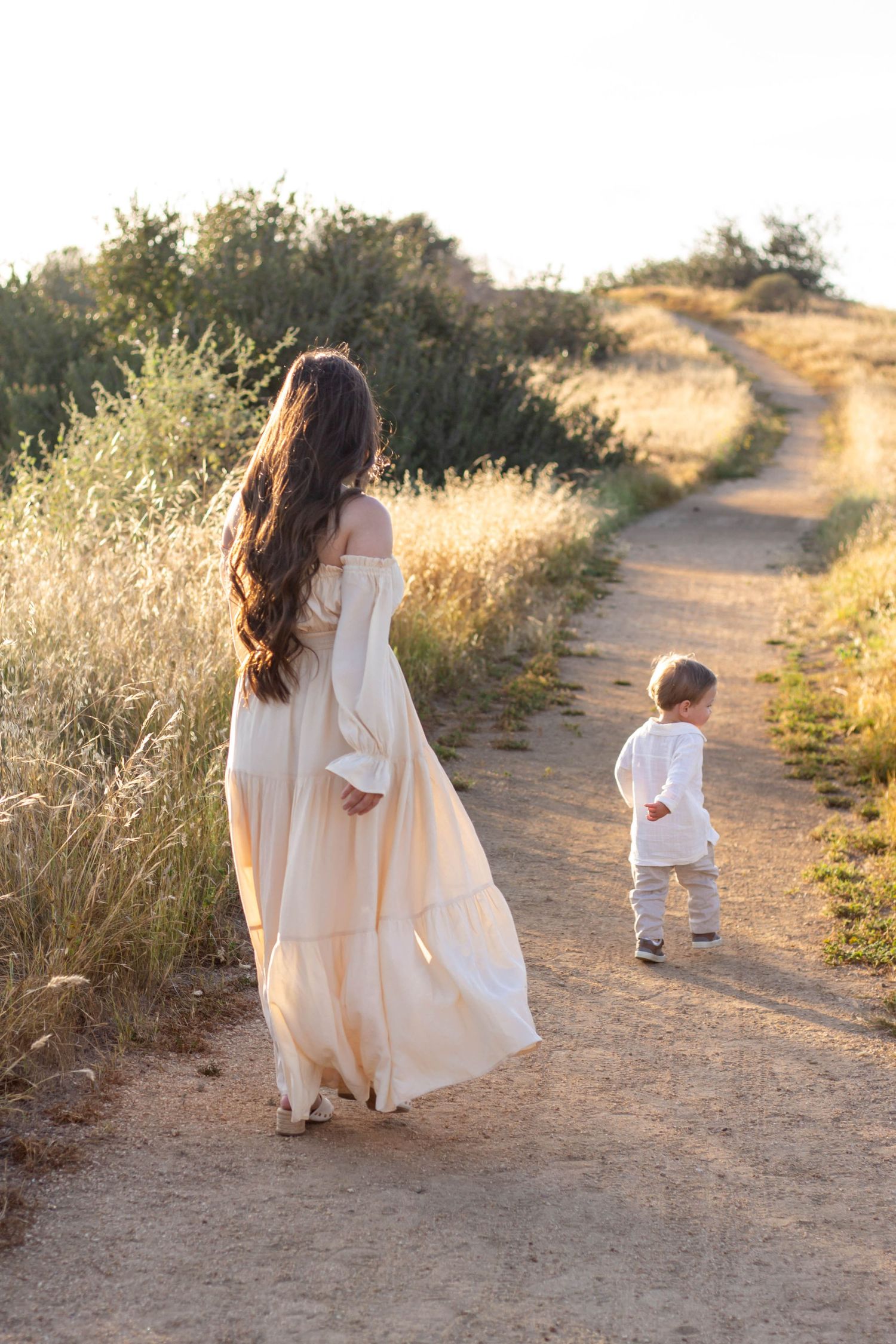 Mother in flowing cream dress walks dirt path with toddler in white shirt at golden sunset.