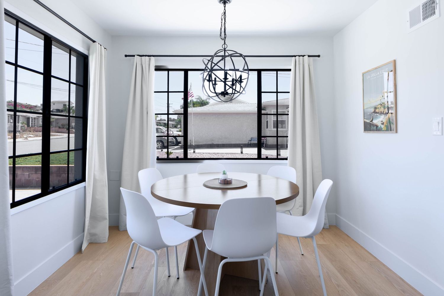 Bright dining room with round wood table, white chairs, black-framed windows, and an orb chandelier over light hardwood floors.