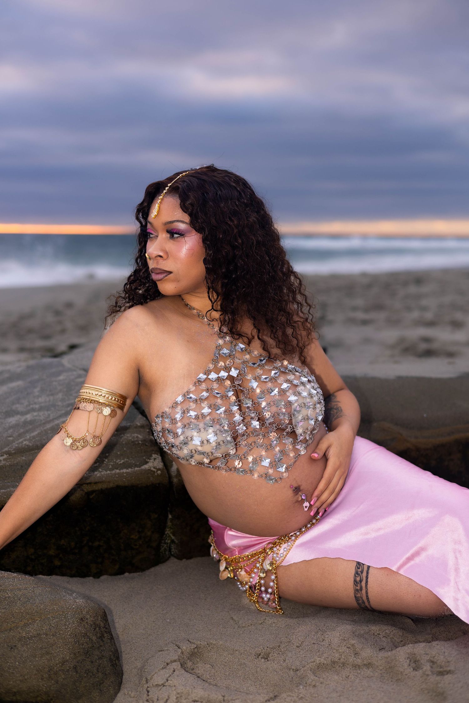 Pregnant woman in silver sequin top and pink skirt poses on beach rocks at sunset with curly hair and gold jewelry.