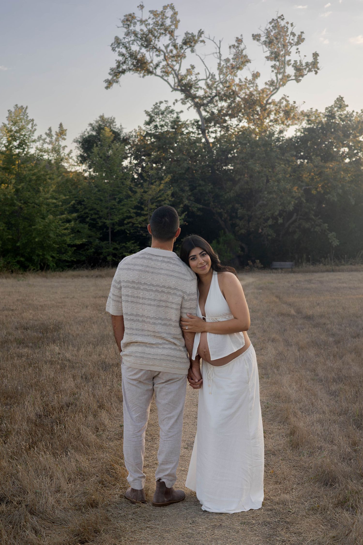 Couple holding hands in a golden field at sunset, woman in white dress smiling, trees in background.