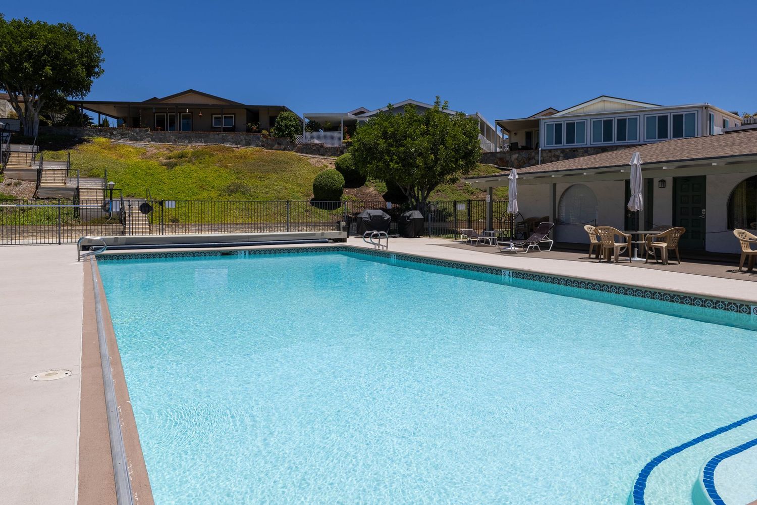 Sparkling outdoor community pool with clear blue water surrounded by a patio, lounge chairs, and hillside homes on a sunny day.
