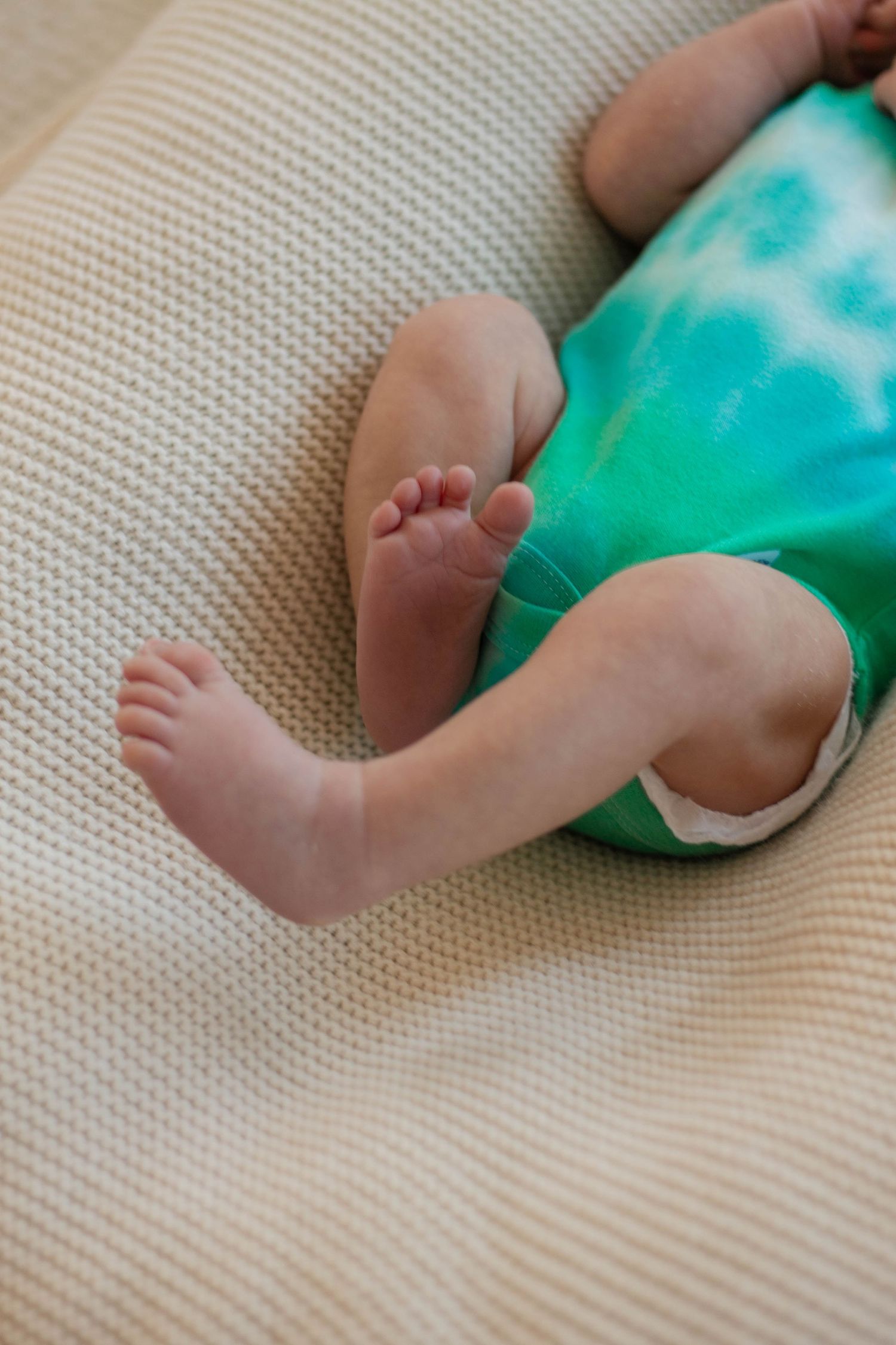 Close-up of baby's bare feet and legs on a textured beige surface wearing a turquoise and white outfit.