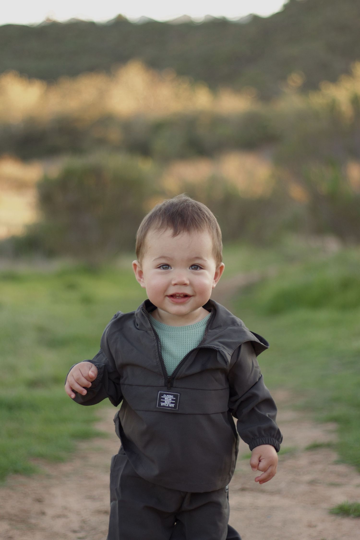 A happy toddler boy in a dark jacket smiles while walking on a nature trail at golden hour.