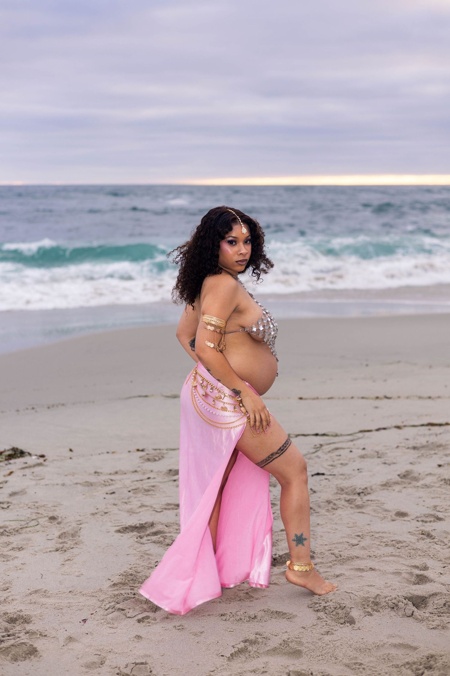 Pregnant woman in pink skirt and embellished top posing on sandy beach at sunset with ocean waves behind her.