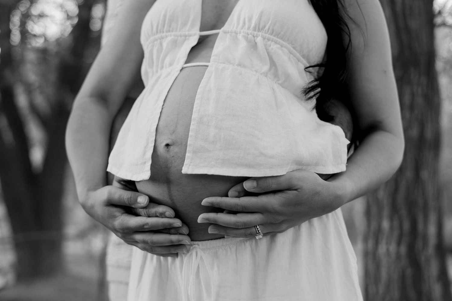 Black and white photo of a pregnant woman in a white dress cradling her baby bump outdoors.