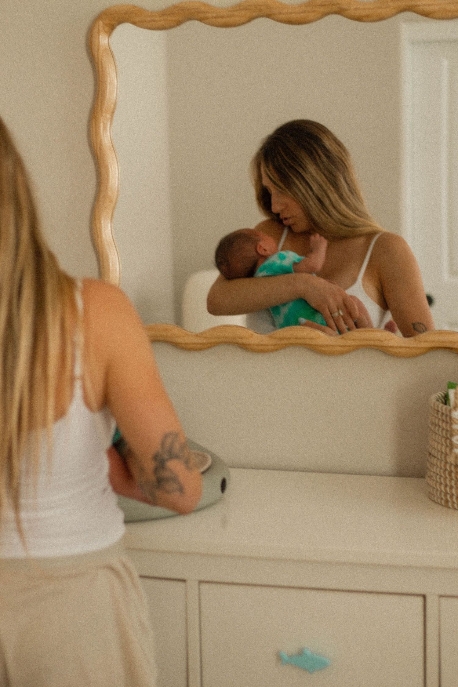 A mother holds her newborn baby while looking at their reflection in a wooden-framed mirror above a white dresser.