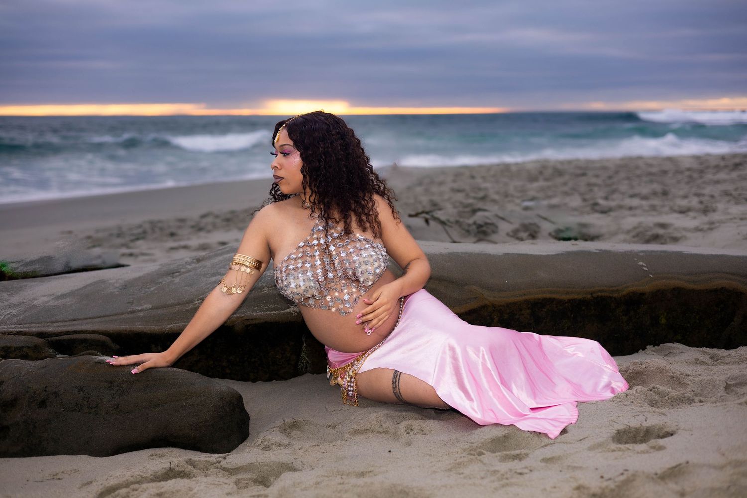 Pregnant woman in pink skirt and beaded top poses on sandy beach at dusk with ocean waves behind her.