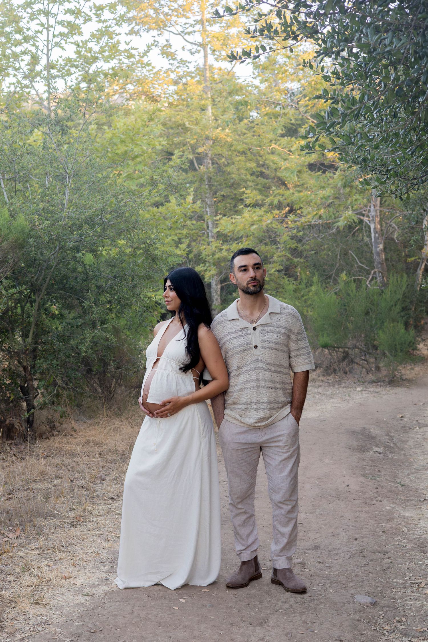Pregnant woman in white maxi dress and man standing on forest path, cradling baby bump outdoors.