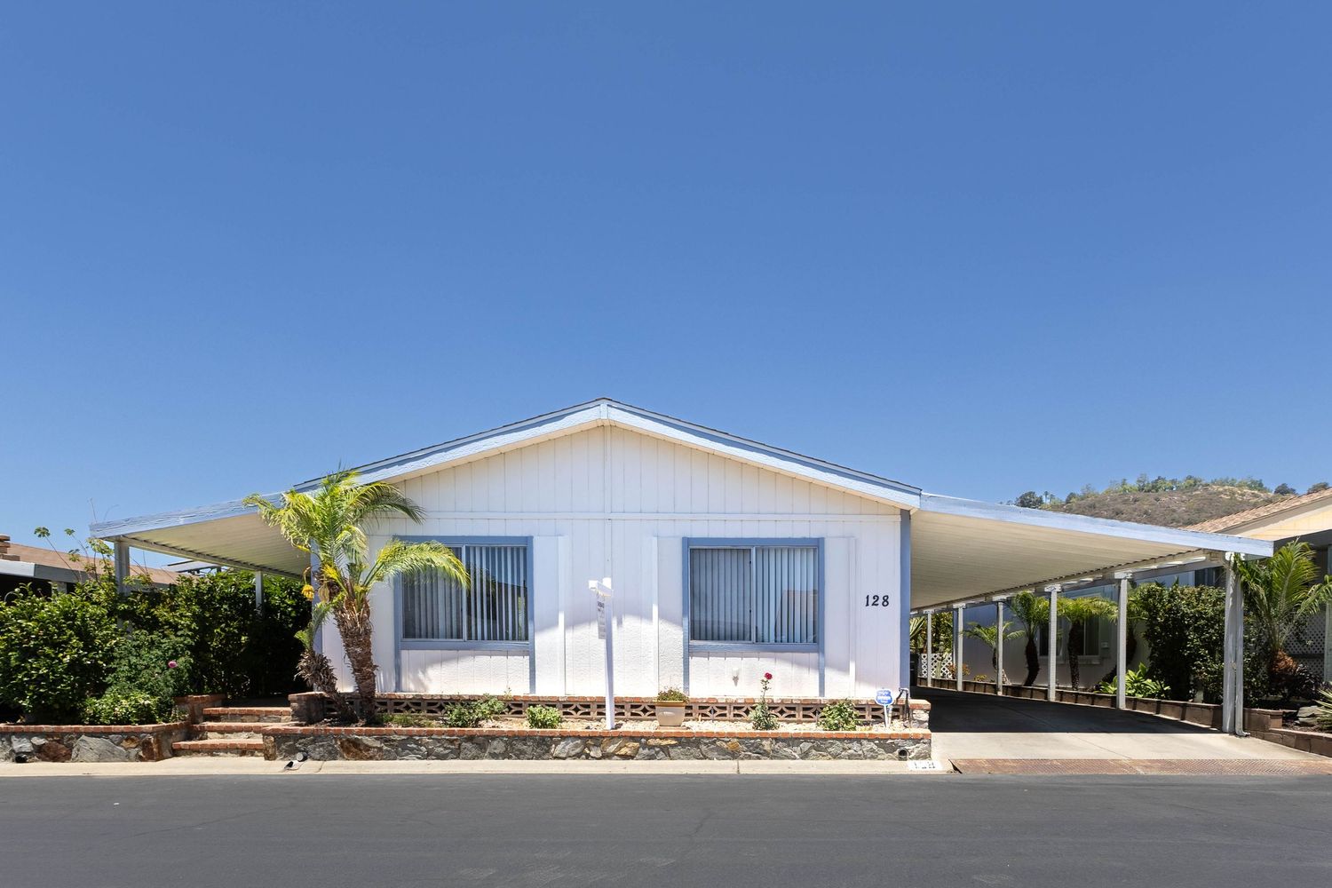 White single-wide mobile home with carport, palm tree, and manicured landscaping under a clear blue sky.