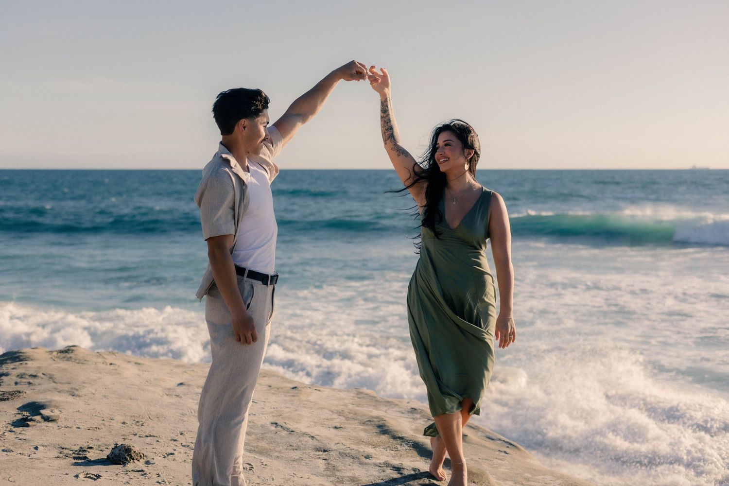 Happy couple dancing on the beach at sunset, man spinning woman in a green dress.