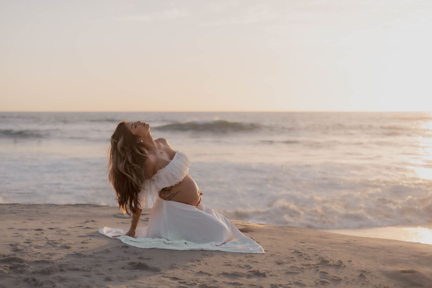 Woman in white outfit practicing yoga on sandy beach at sunset with ocean waves in background.