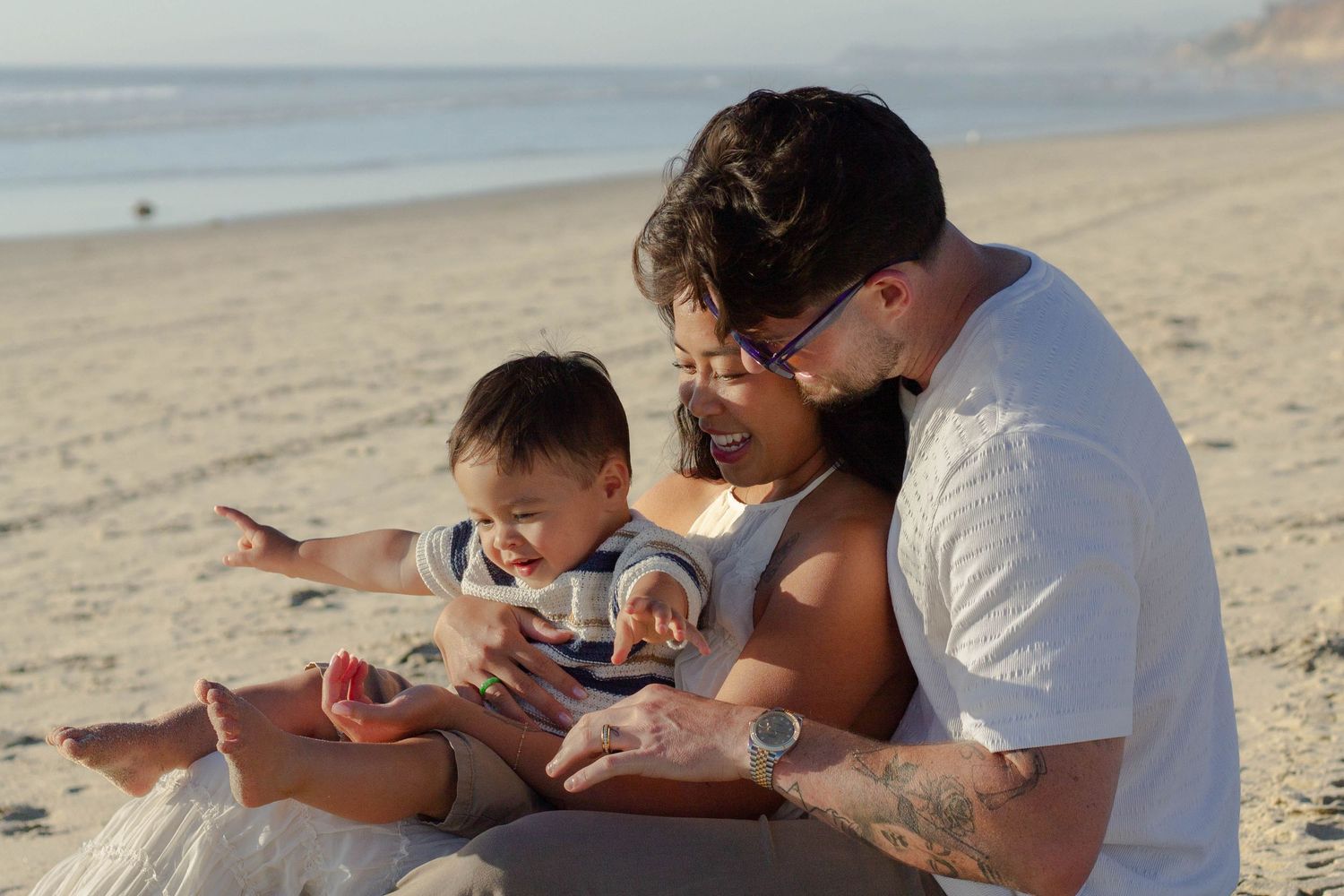A young family sits together on a sunny beach, with their baby pointing at something in dad's hand.