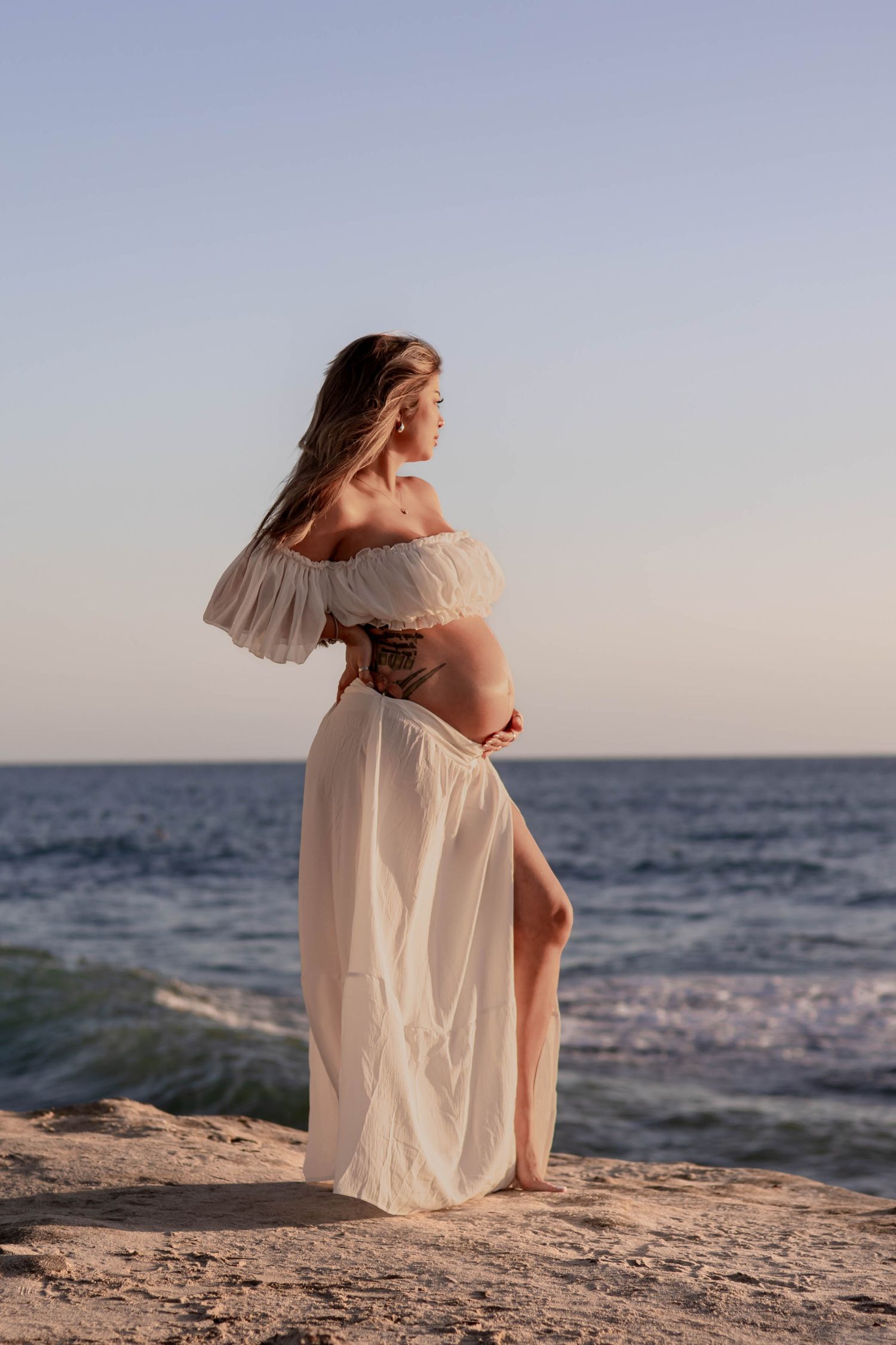 Pregnant woman in white off-shoulder dress cradling her belly on rocky beach at sunset.
