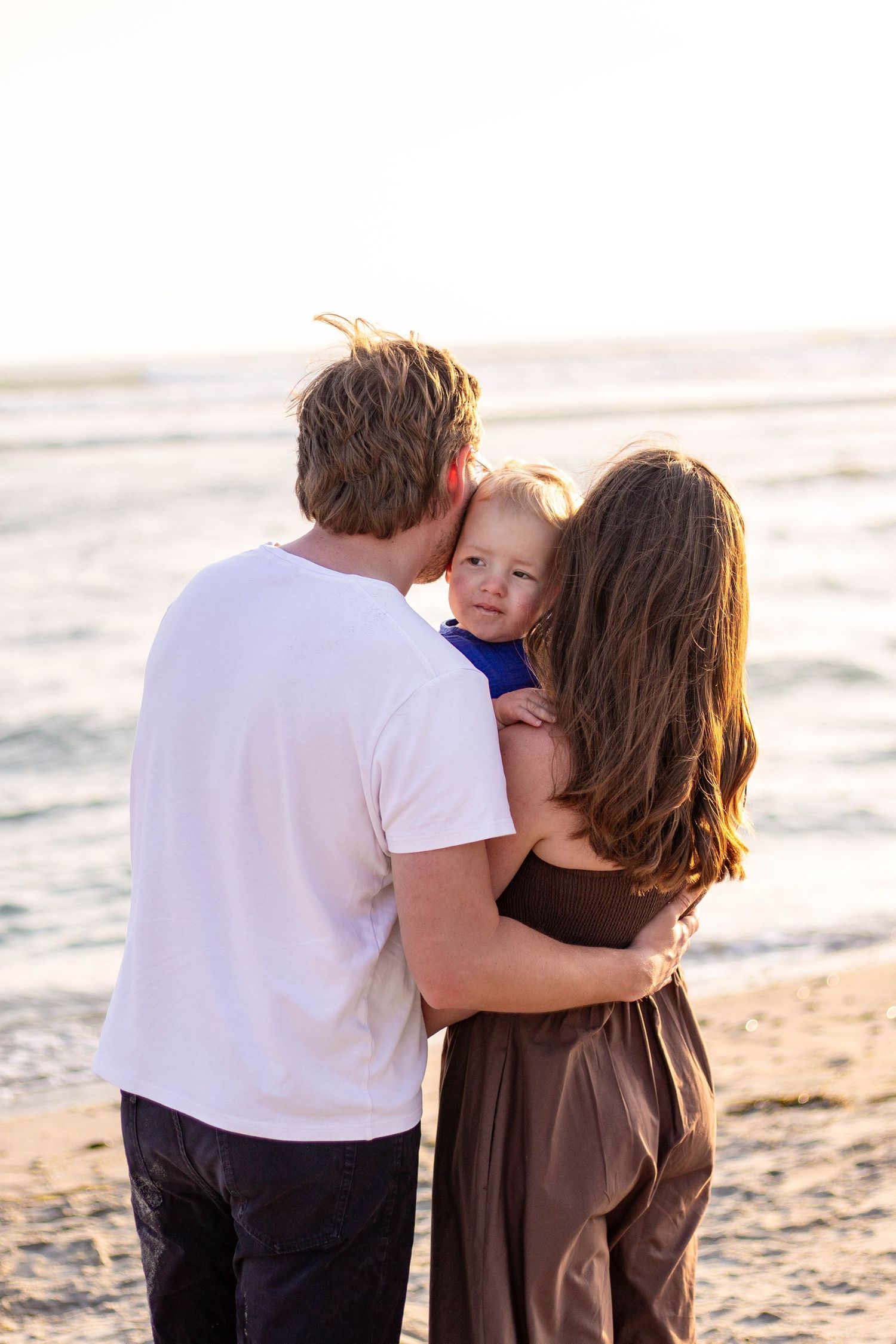 Parents at beach sunset kissing and embracing toddler child near ocean shoreline.