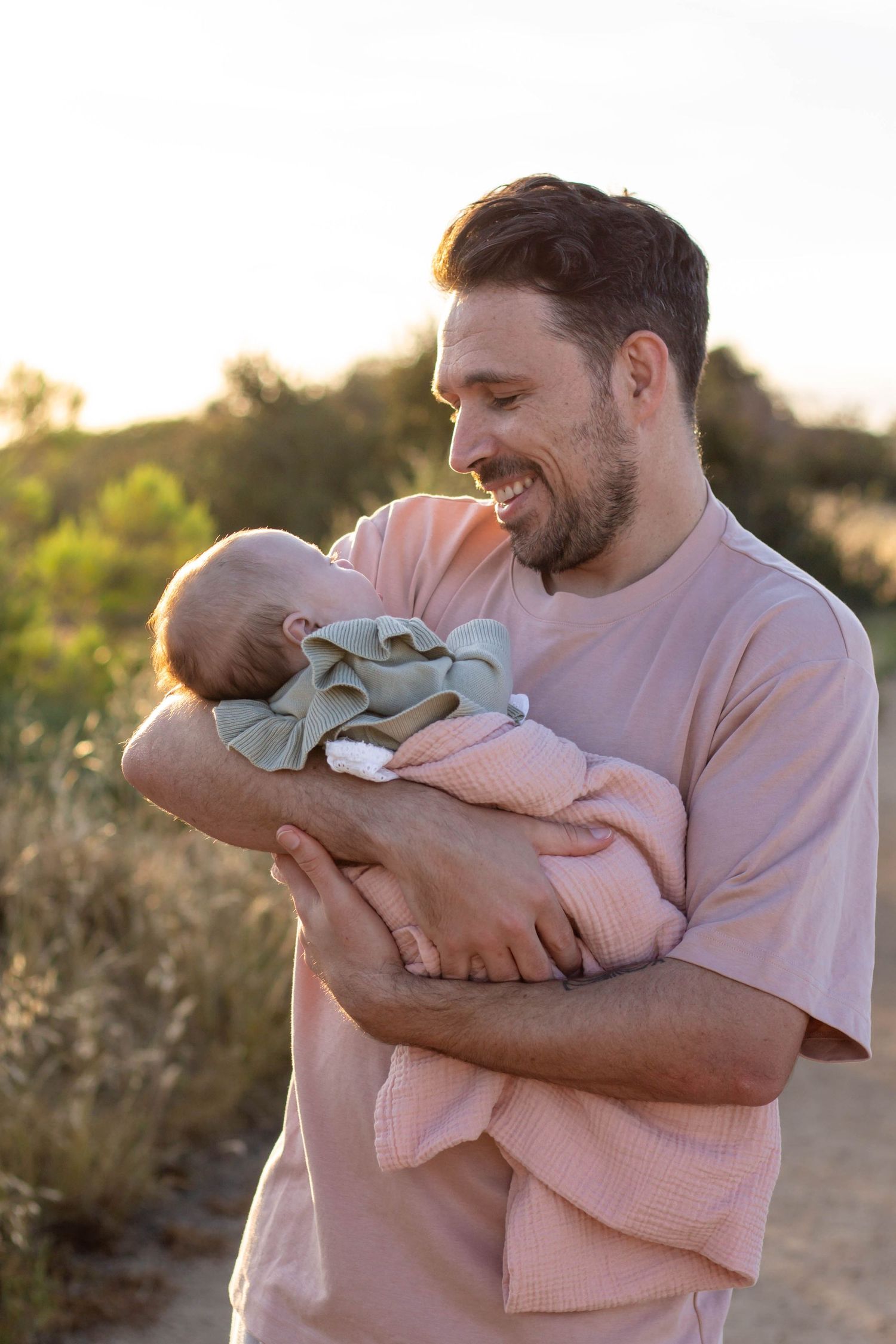 Smiling father cradling newborn baby outdoors during warm golden sunset.