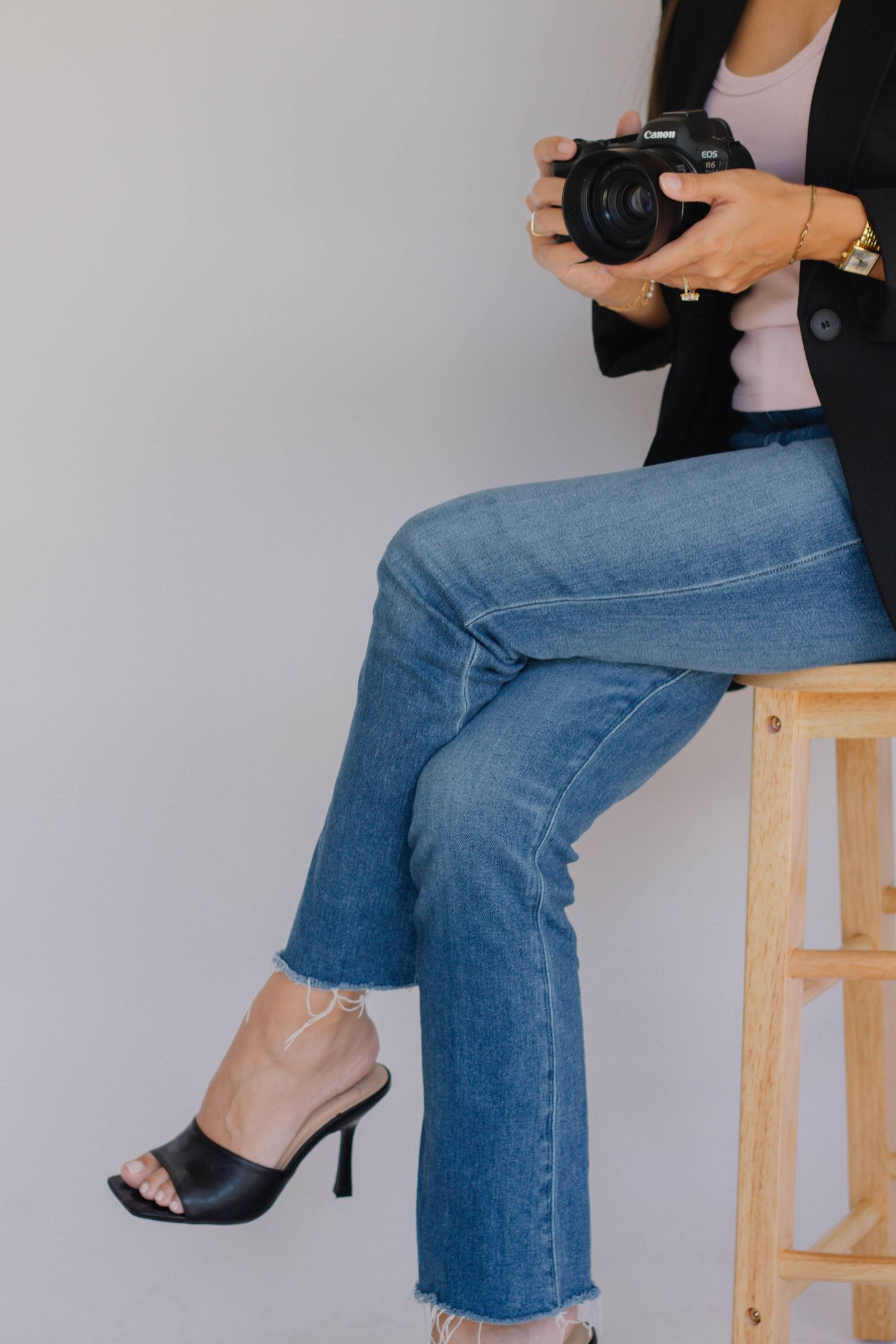Woman sitting on wooden stool holding Canon DSLR camera, wearing blue jeans, black blazer, and black heeled mules.